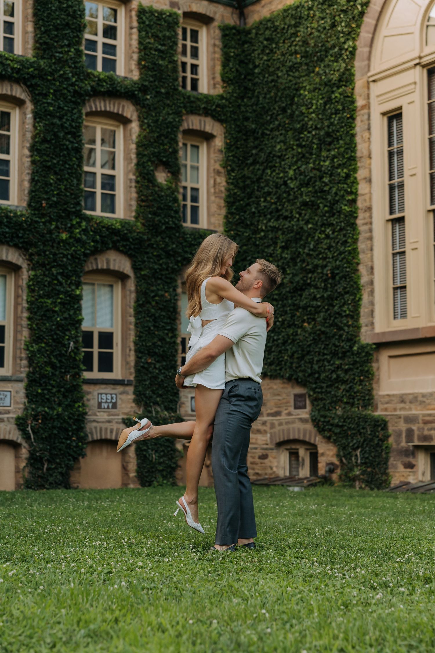 Couple embracing on green lawn in front of ivy-covered brick building, man lifting woman in romantic outdoor setting.