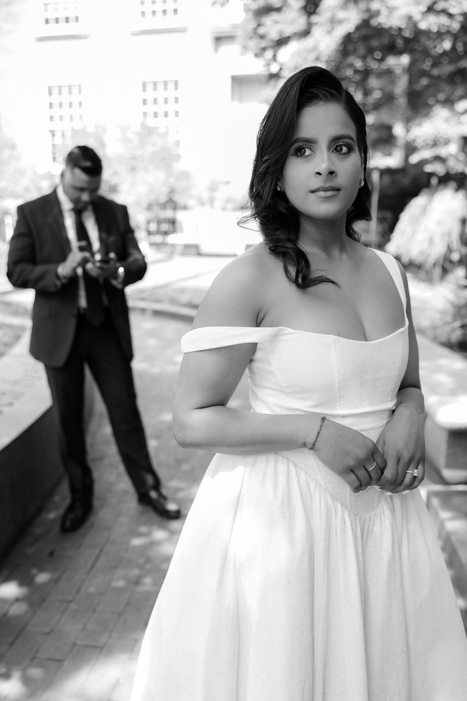 Black and white portrait of elegant bride in off-shoulder gown with groom in background.
