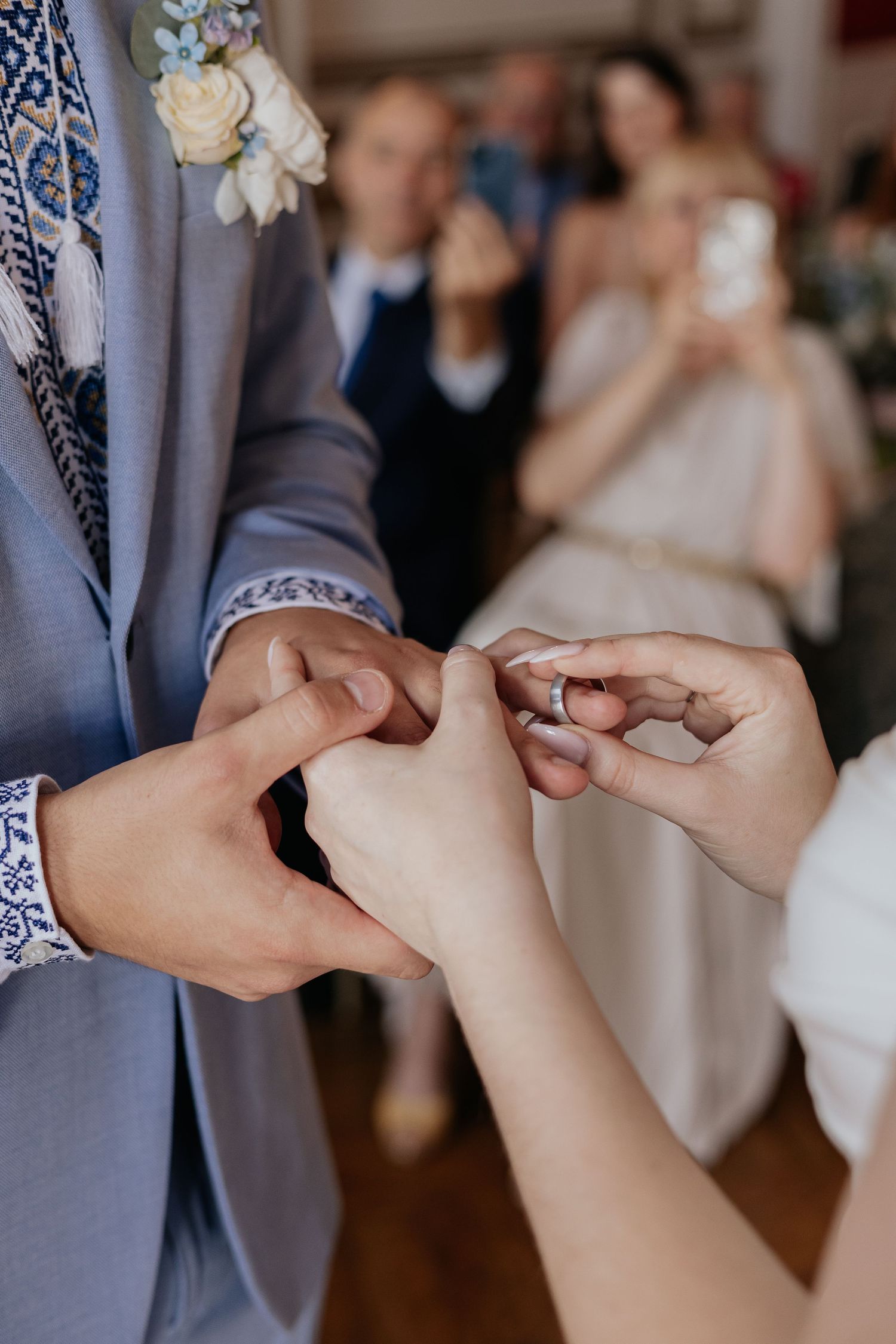 Bride sliding wedding ring onto groom's finger during indoor ceremony with guests in background.