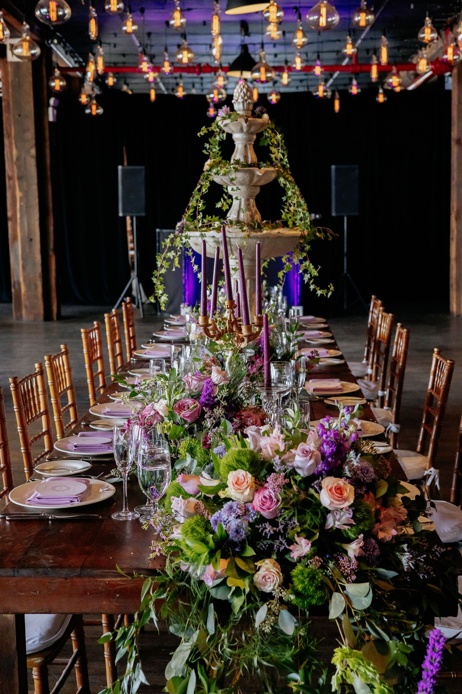 Long farm table with lush floral garland, purple candles, and ornate fountain centerpiece in dark venue.