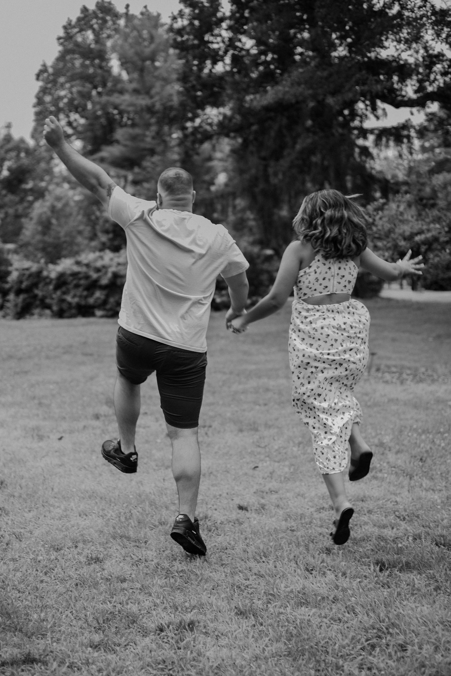 Black and white photo of a joyful couple running hand in hand across a grassy park.
