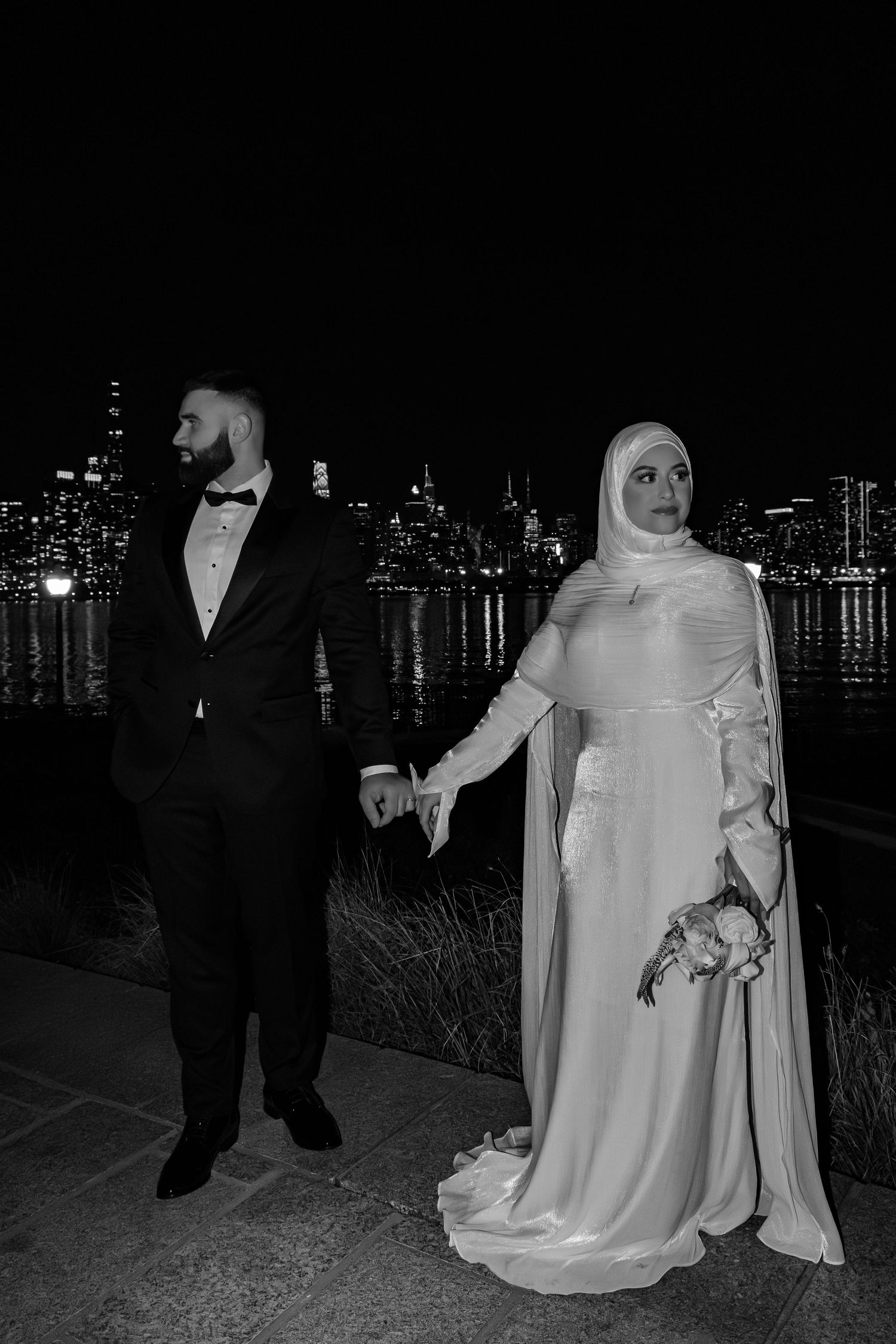 Black and white photo of Muslim wedding couple standing by waterfront with city skyline backdrop.