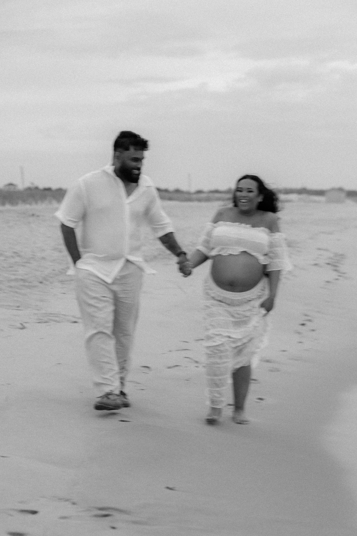 Pregnant couple holding hands and walking on sandy beach, smiling during outdoor maternity photography session.
