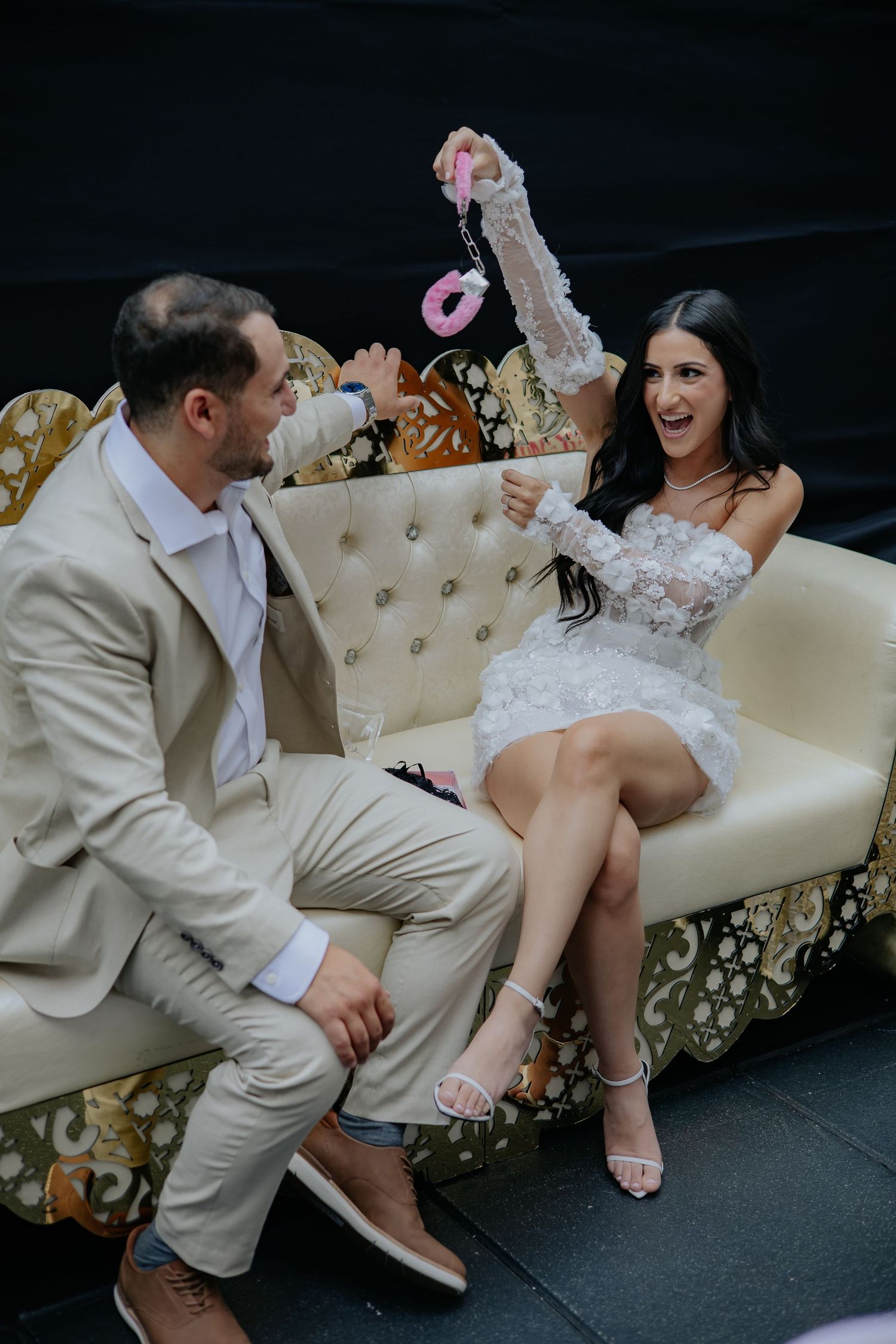 Bride in white lace mini dress laughing with groom in beige suit on ornate gold sofa at wedding reception.