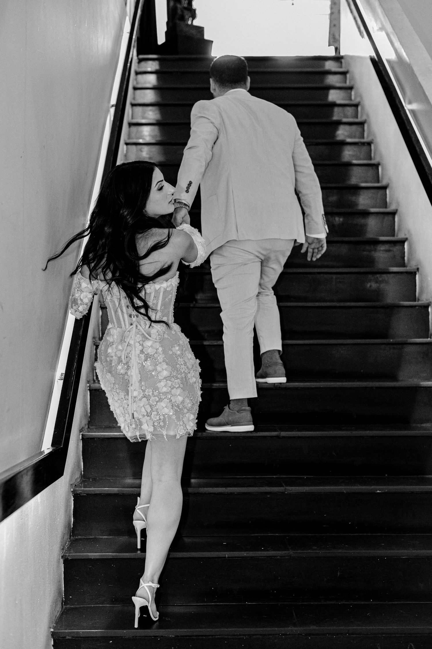 Black and white photo of bride kissing groom's hand as they walk up a staircase at their wedding venue.