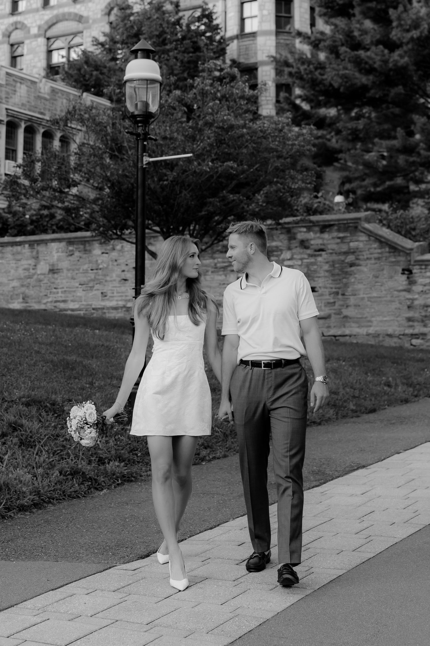 Black and white photo of a couple walking on a path, woman in white dress holding flowers, man in dress pants and polo shirt.