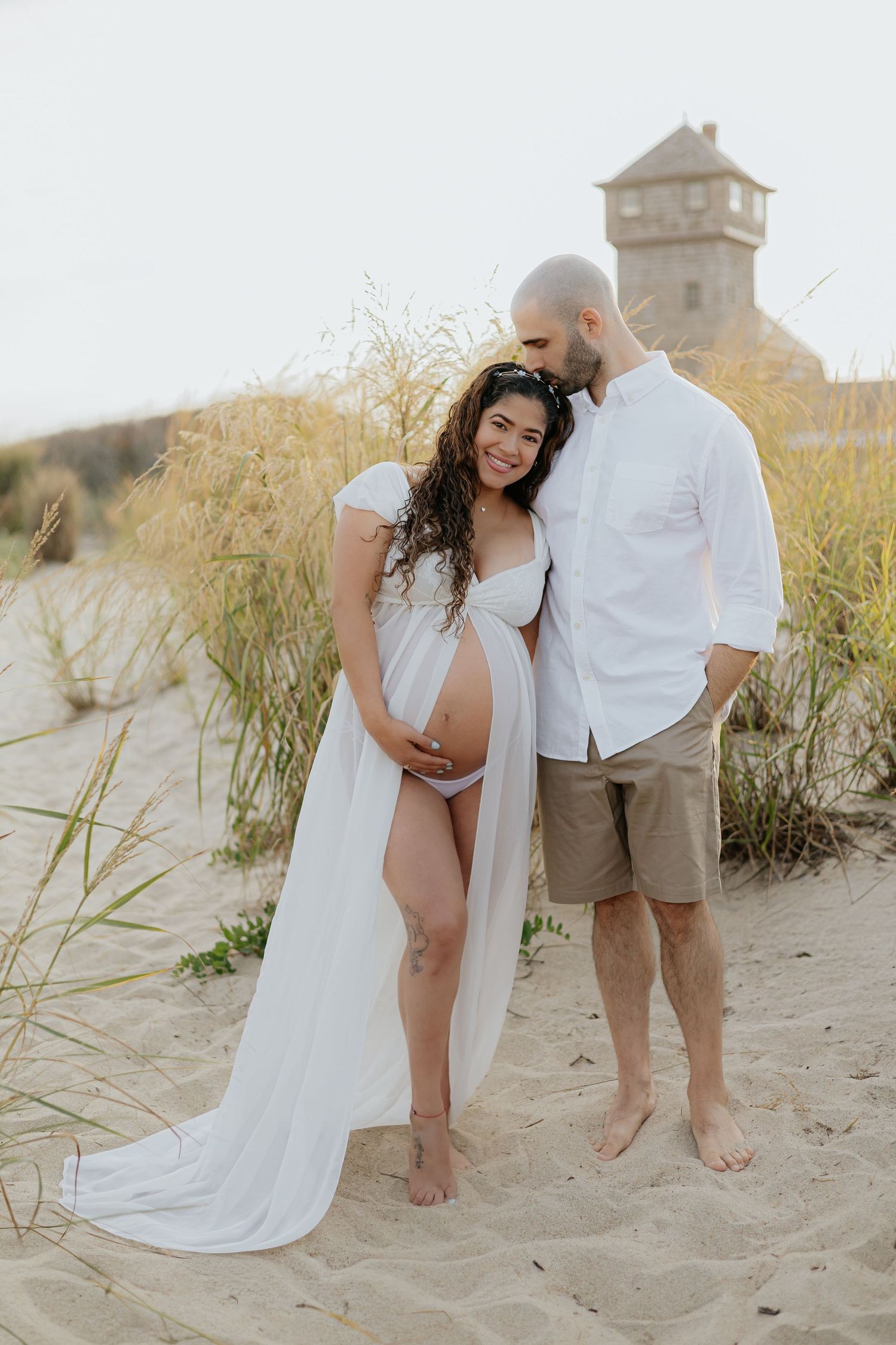 Pregnant woman in white flowing dress with partner on sandy beach dunes during maternity photoshoot.