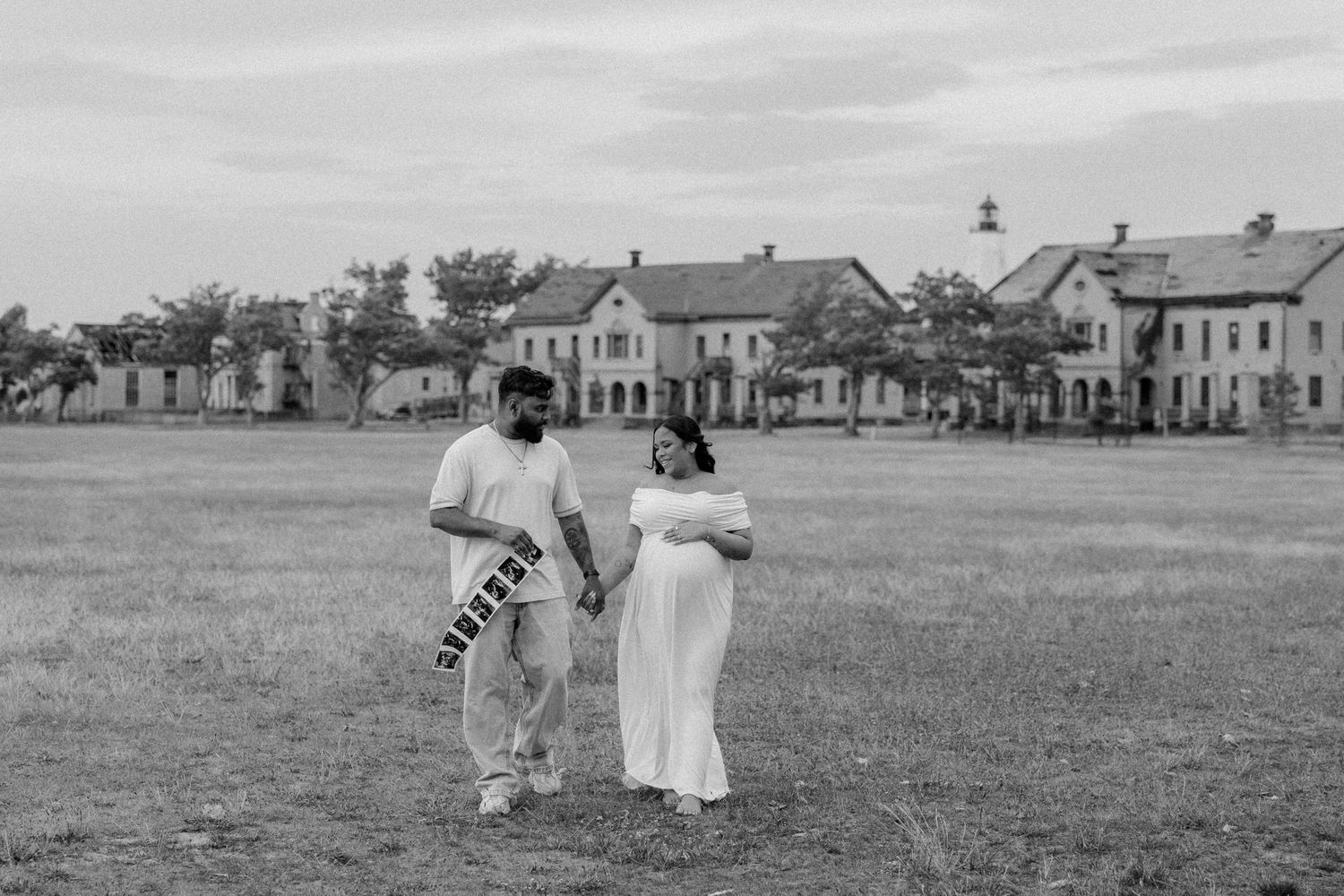 Black and white maternity photo of a couple holding hands in a field with historic buildings in the background.