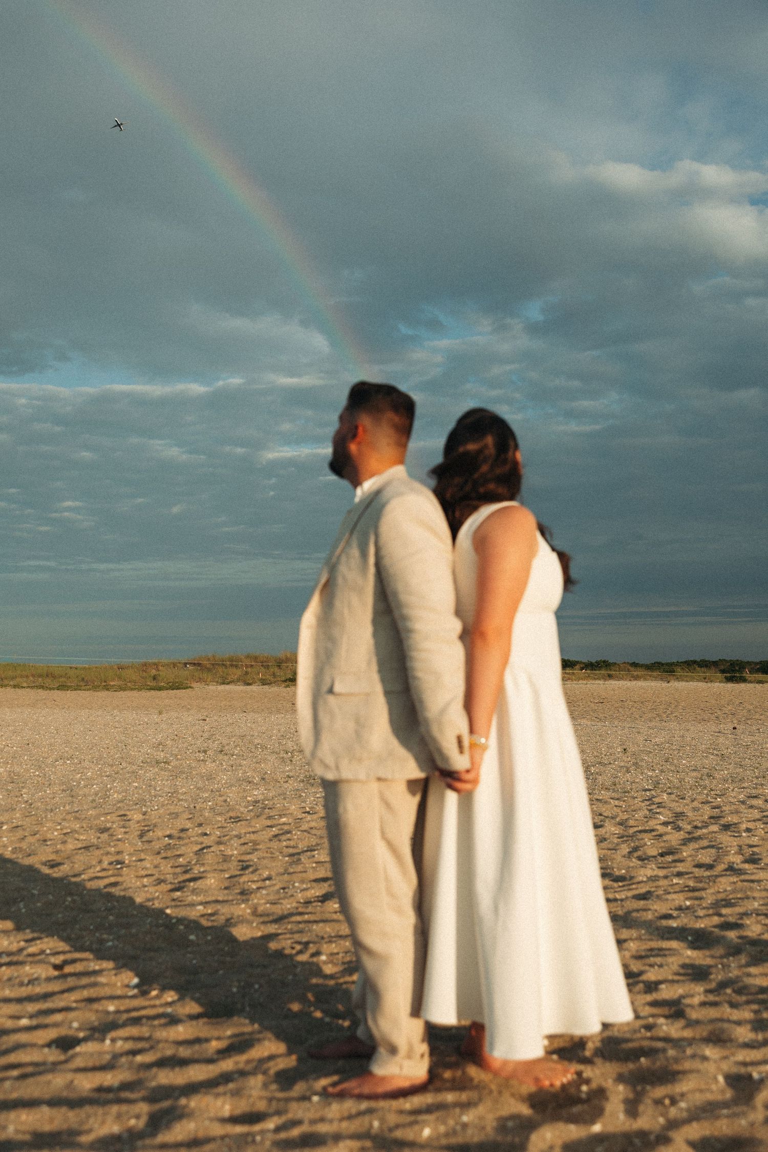 Couple holding hands on beach under rainbow with dramatic cloudy sky at sunset.
