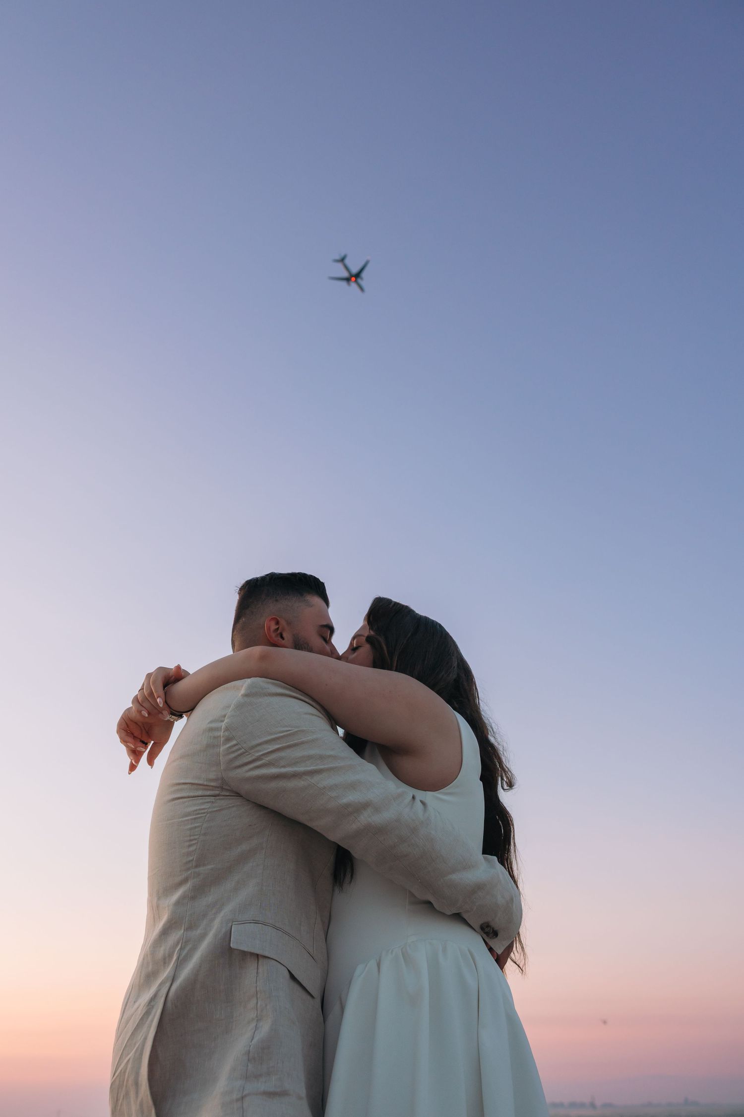 Couple embracing and kissing at sunset with an airplane flying overhead in a pastel blue and pink sky.
