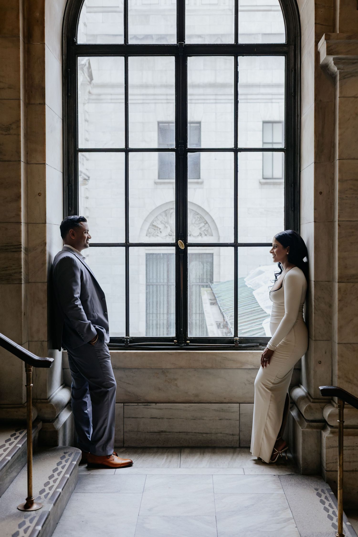 Elegant couple standing by a grand window in a historic marble building.
