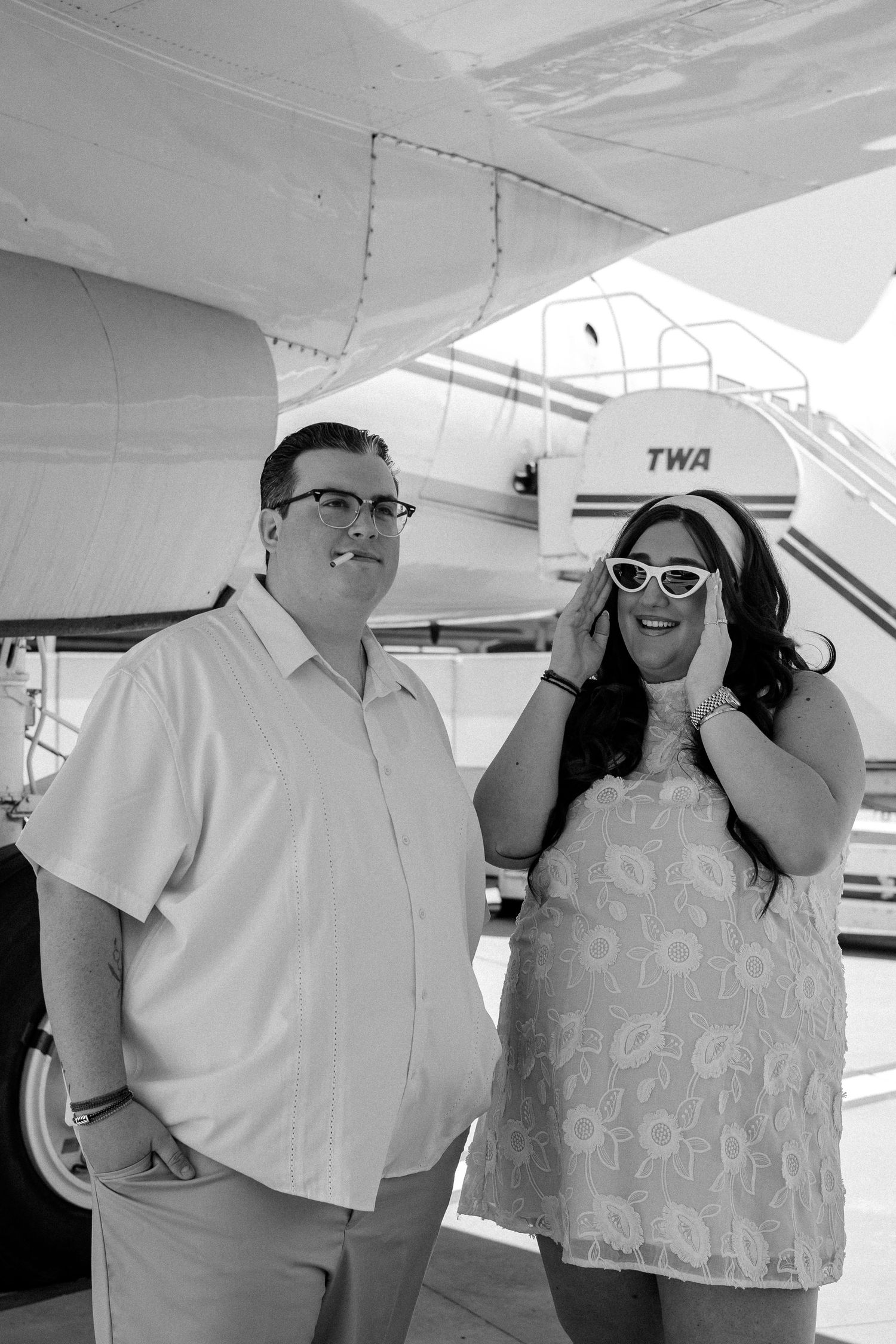 Black and white photo of smiling couple posing in front of a TWA airplane, woman wearing retro sunglasses.