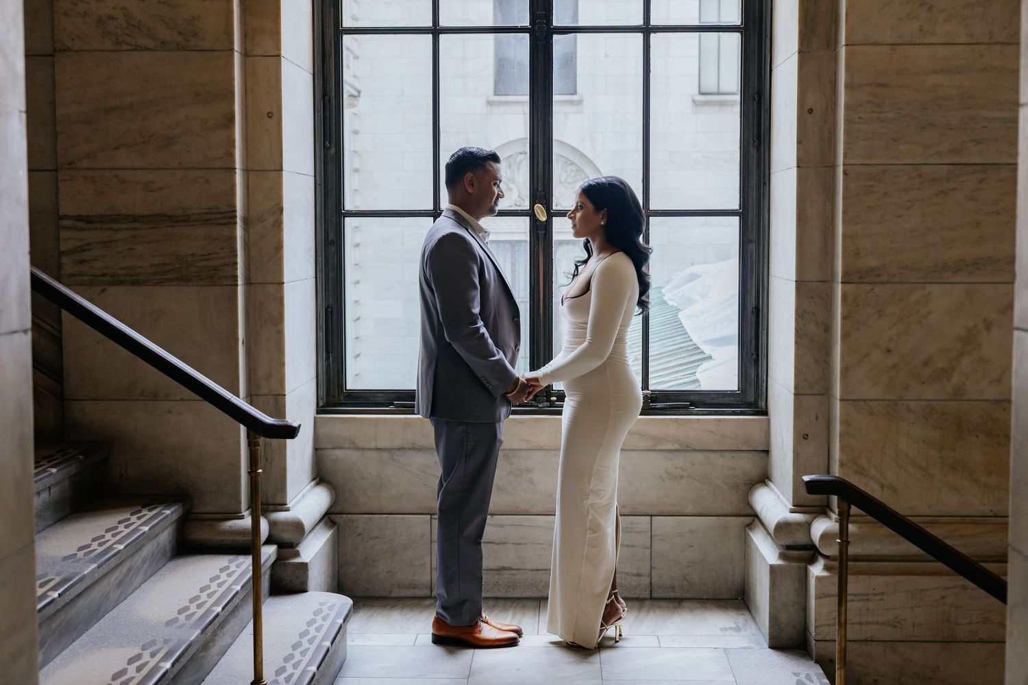 Couple in wedding attire holding hands and facing each other by large windows on marble staircase landing.