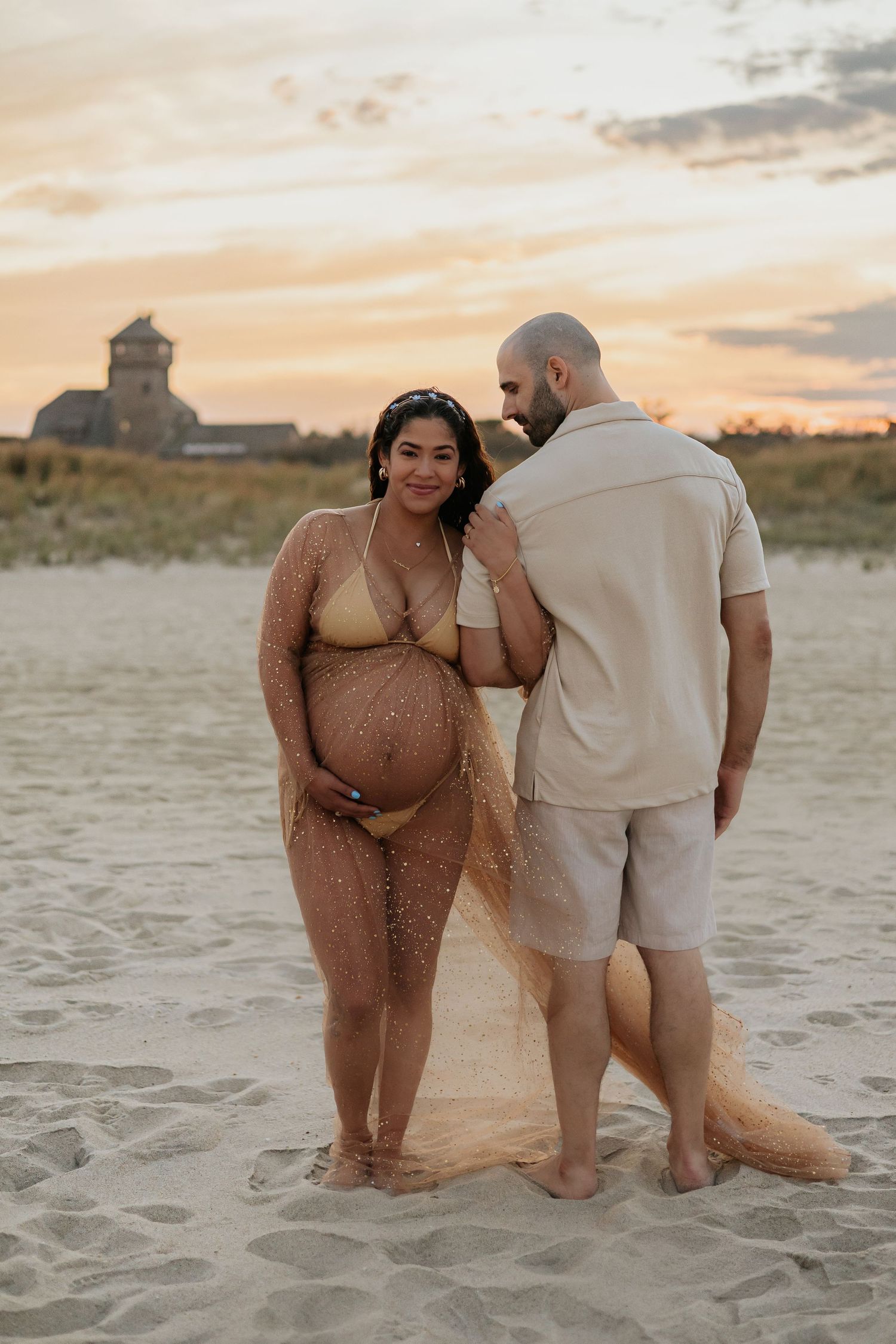 Pregnant woman in gold glitter tulle gown poses with partner on beach at golden hour.