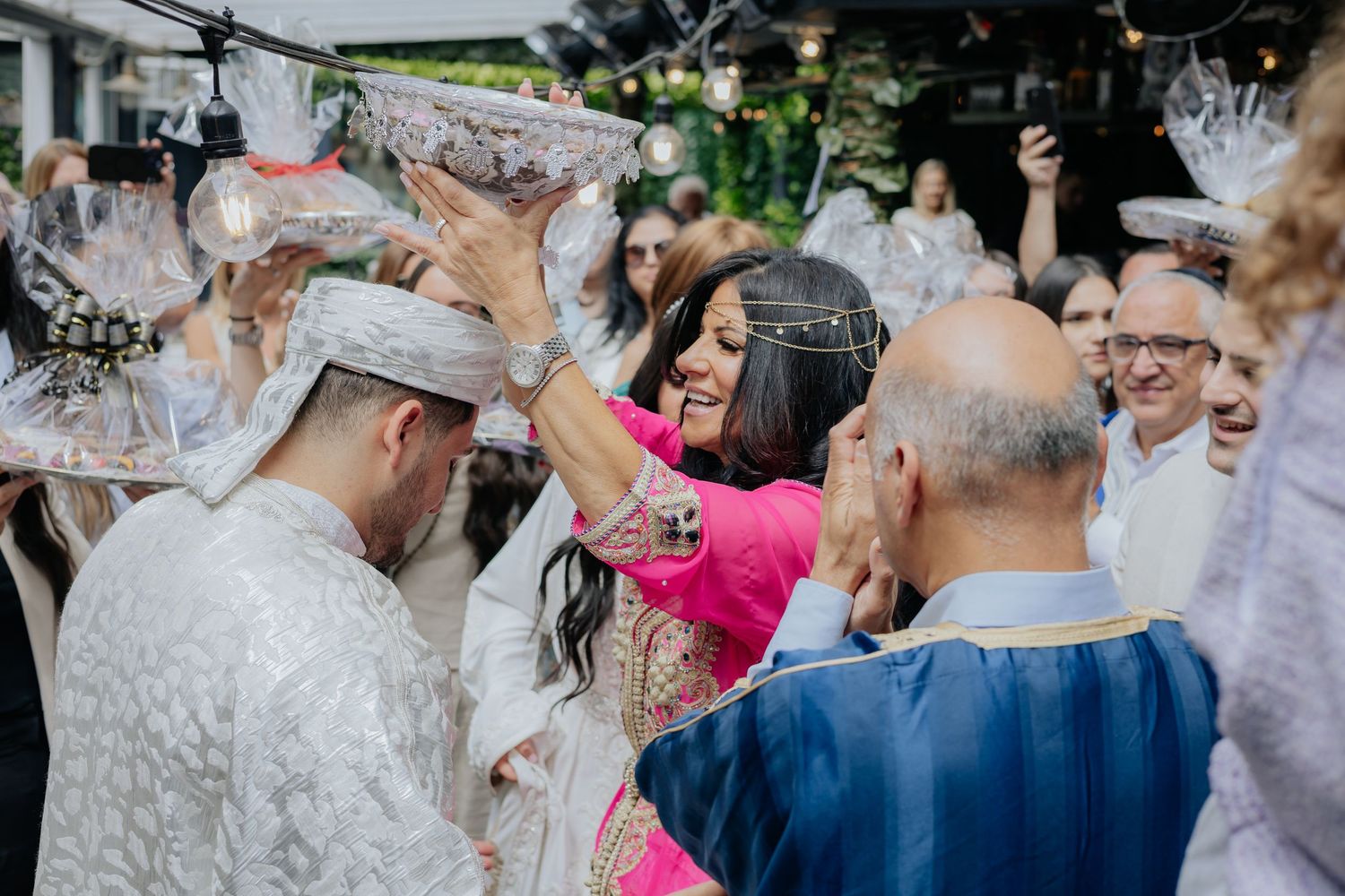 Woman in pink kaftan performs traditional Moroccan wedding ritual with decorated trays over groom and guests outdoors.