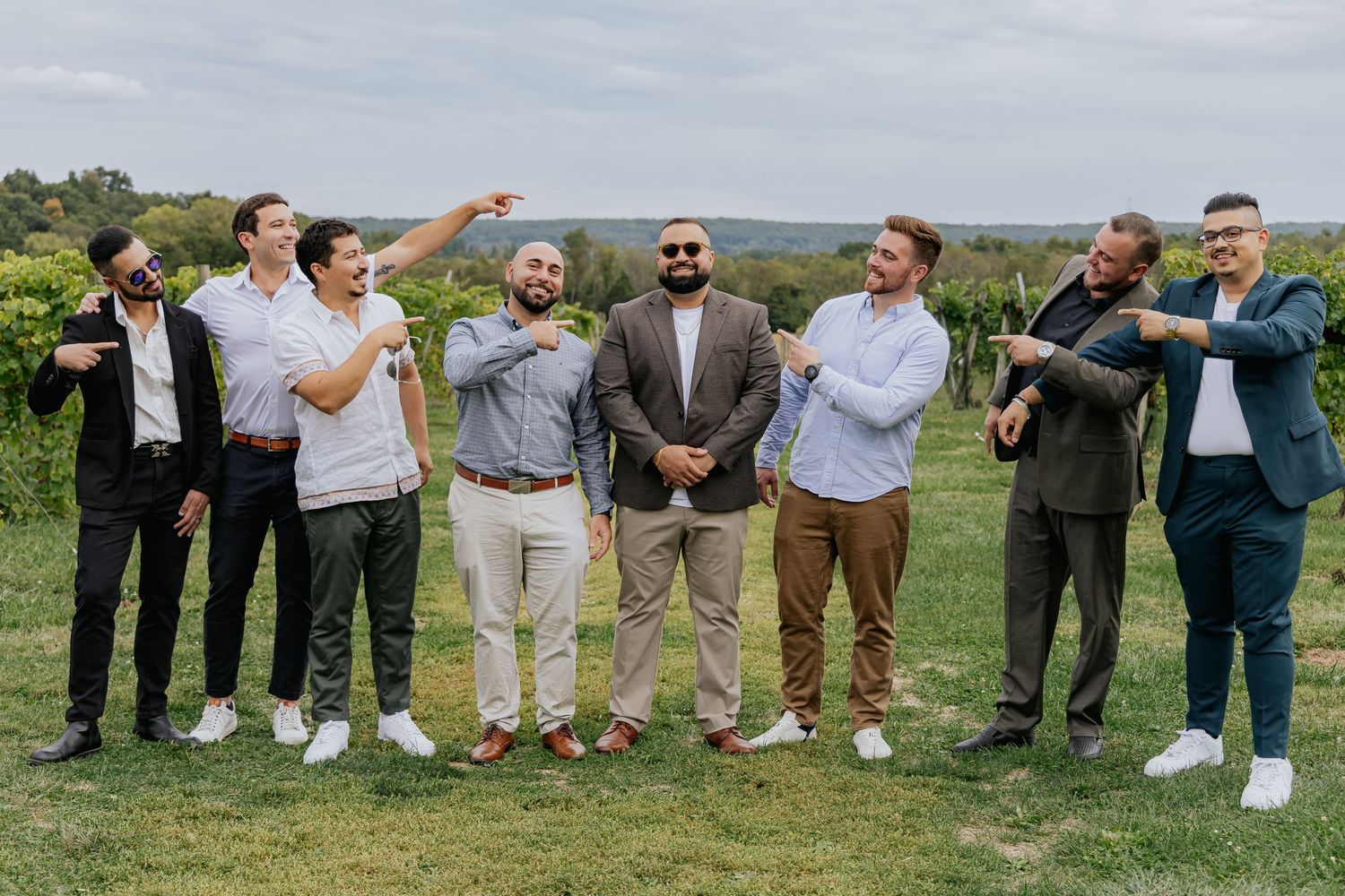 Seven men in formal attire posing playfully and pointing at the groom at a vineyard wedding.