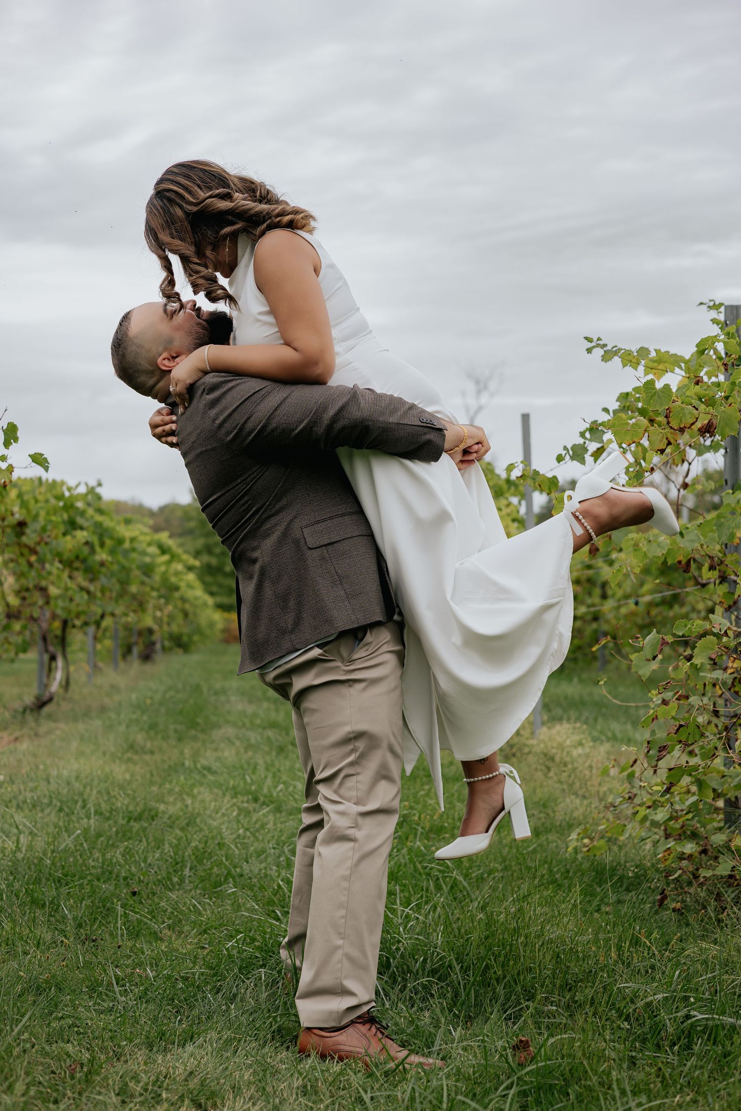 Groom lifting bride in white dress for a kiss among green vineyard rows on cloudy day.