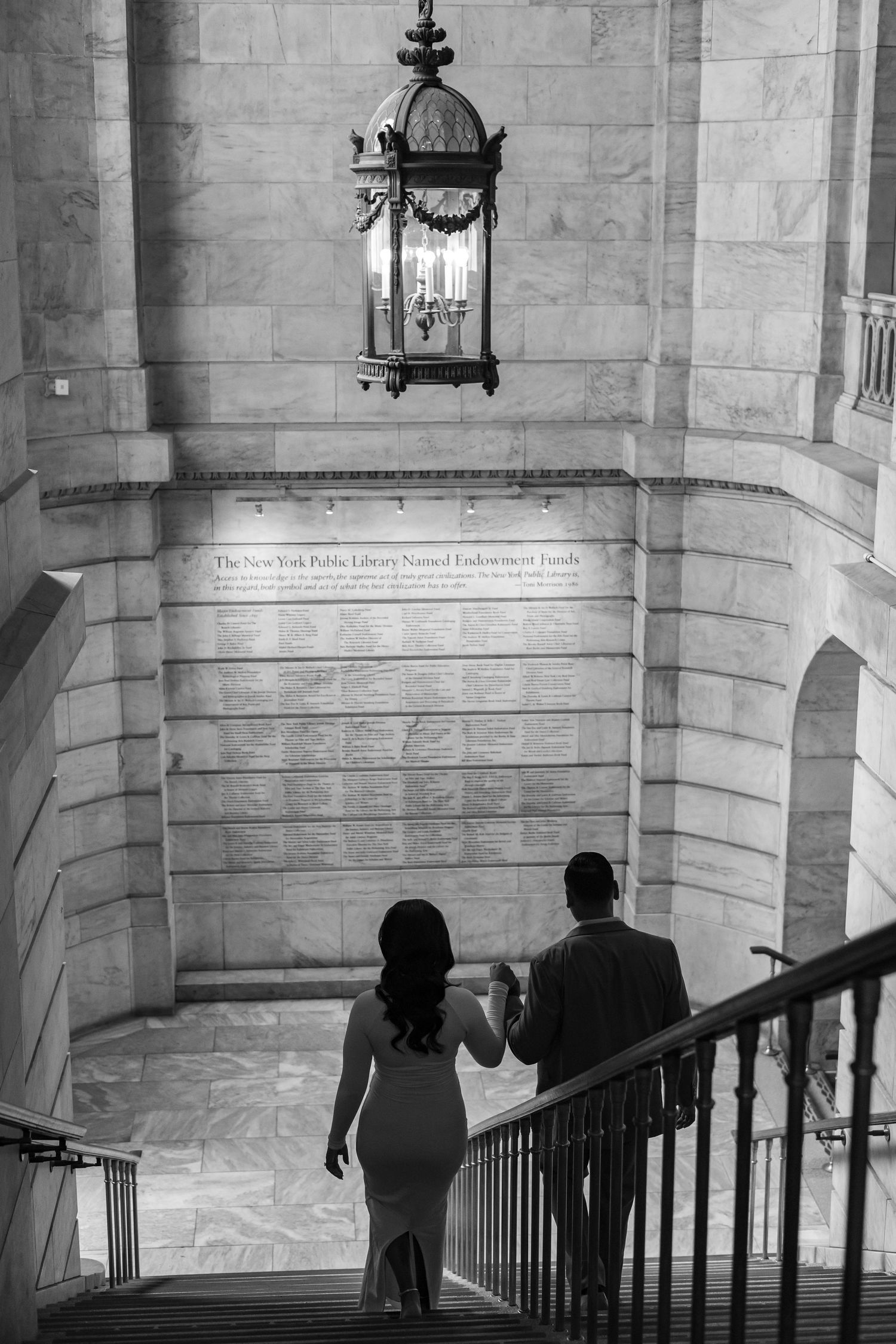 Black and white photo of couple ascending stairs beneath vintage lantern at NYPL.
