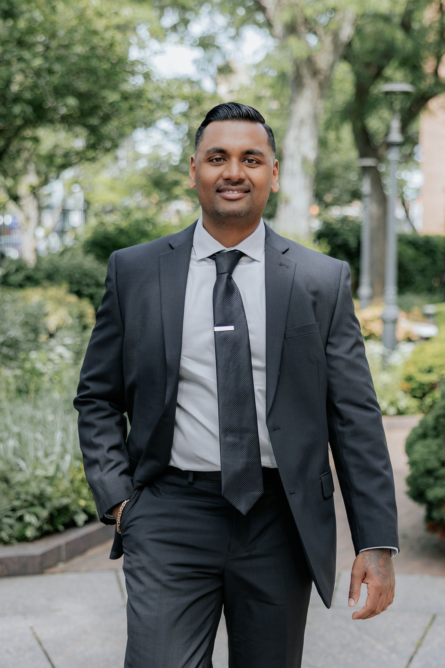 Confident South Asian groom in dark suit and tie smiling while walking in outdoor garden setting.
