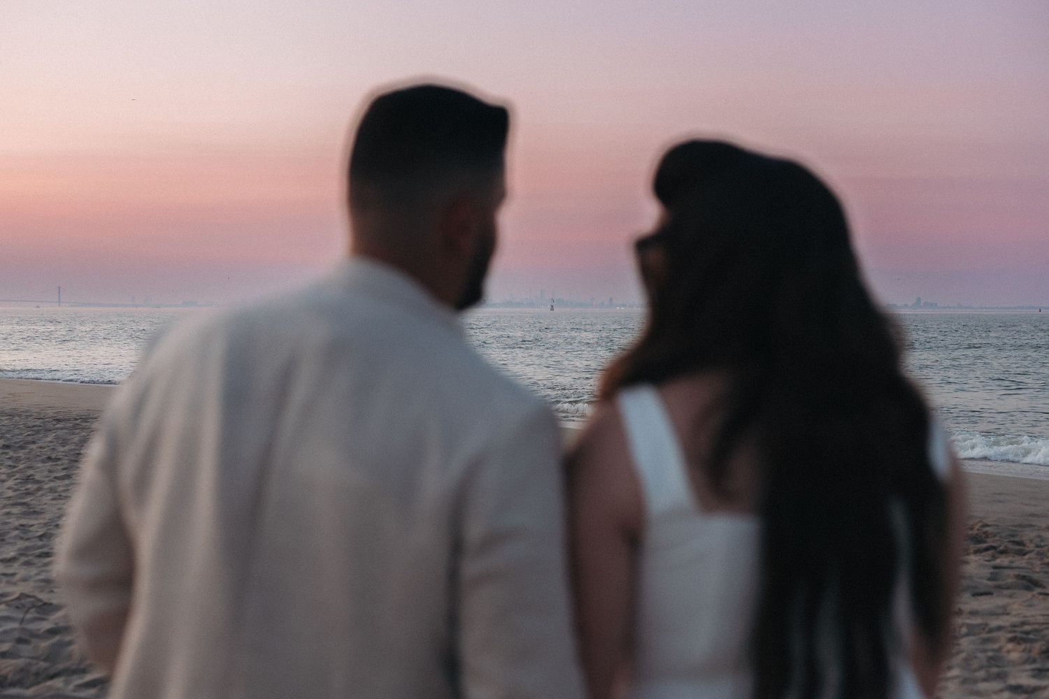 Couple standing on beach at sunset, man in white linen shirt and woman in white dress, pink sky over ocean.