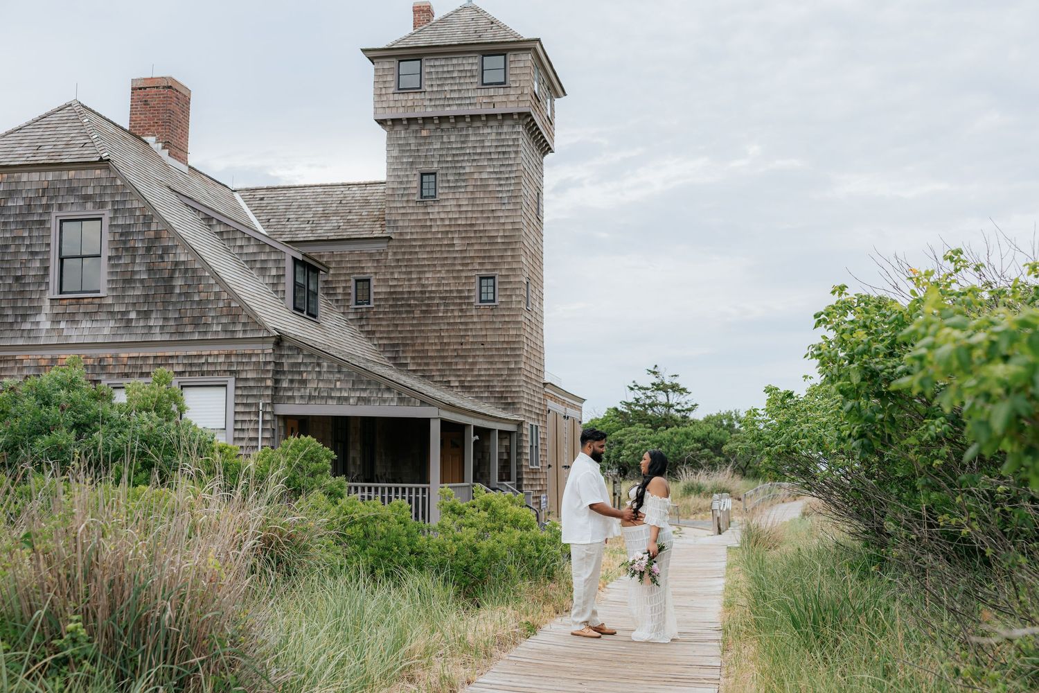 Couple walks on boardwalk path near rustic shingle-style beach house with tower in coastal setting.