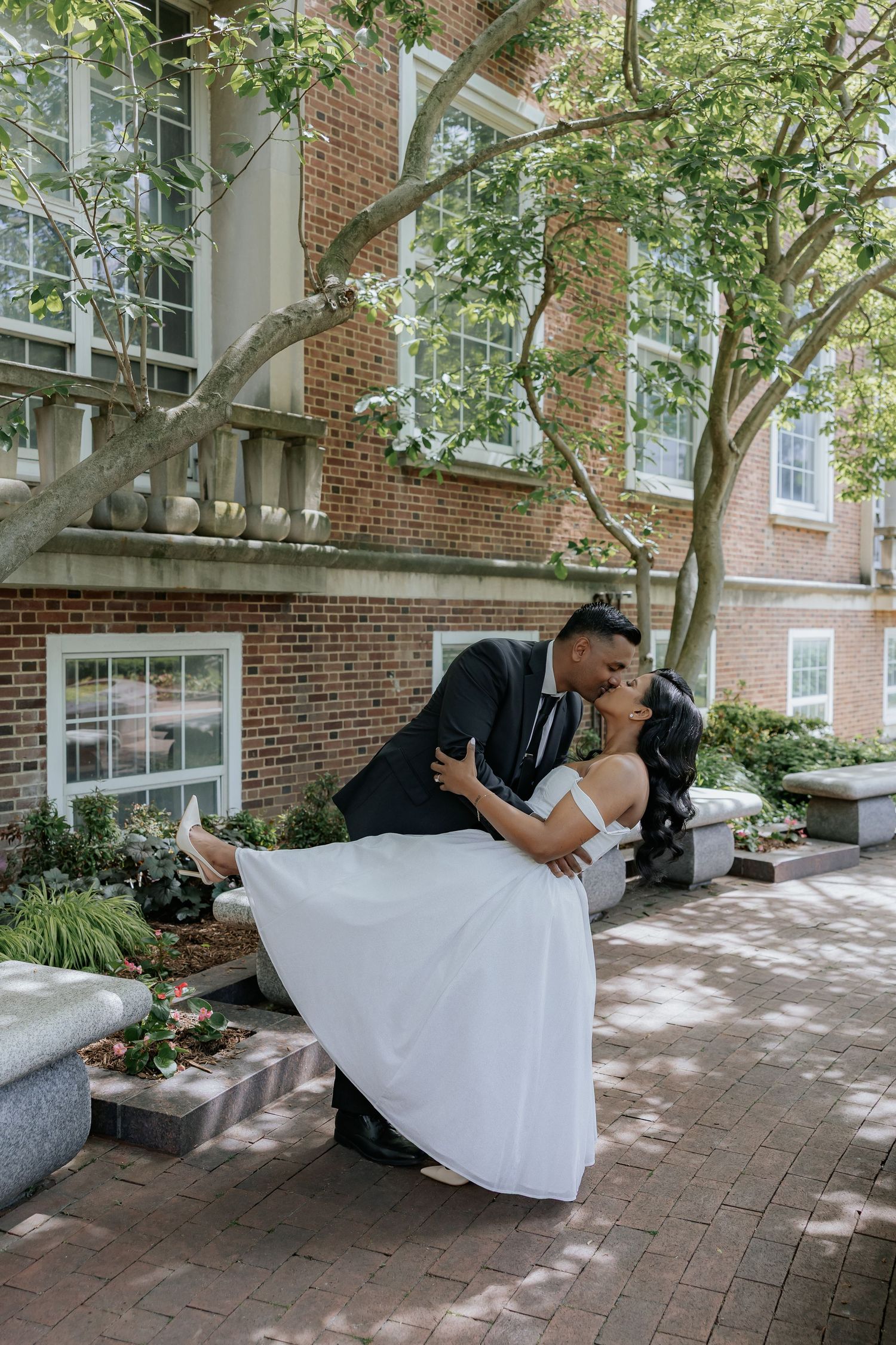 Groom dips bride for a kiss in a lush courtyard outside a historic brick building.