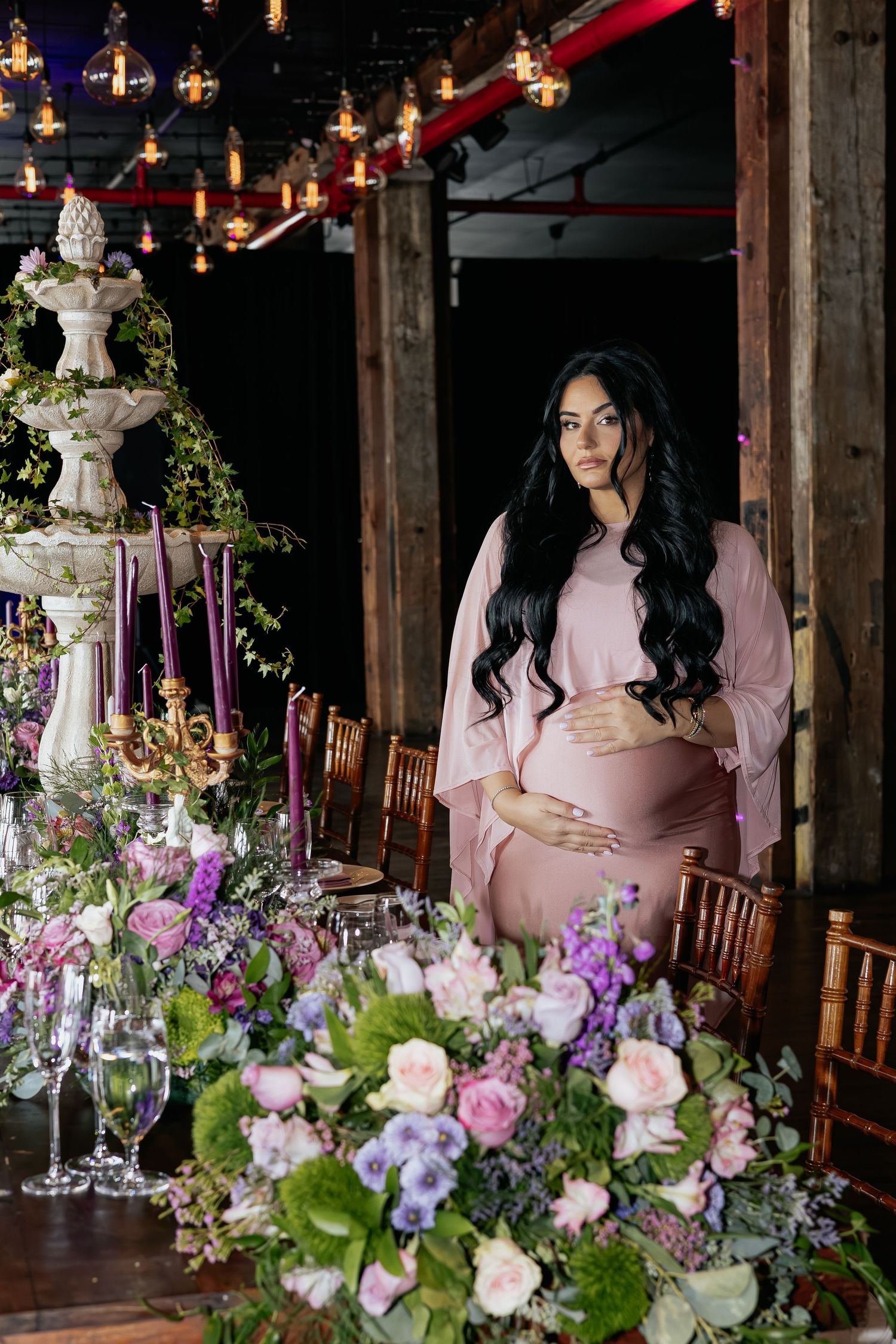 Pregnant woman in blush pink gown posing beside floral baby shower tablescape with fountain.