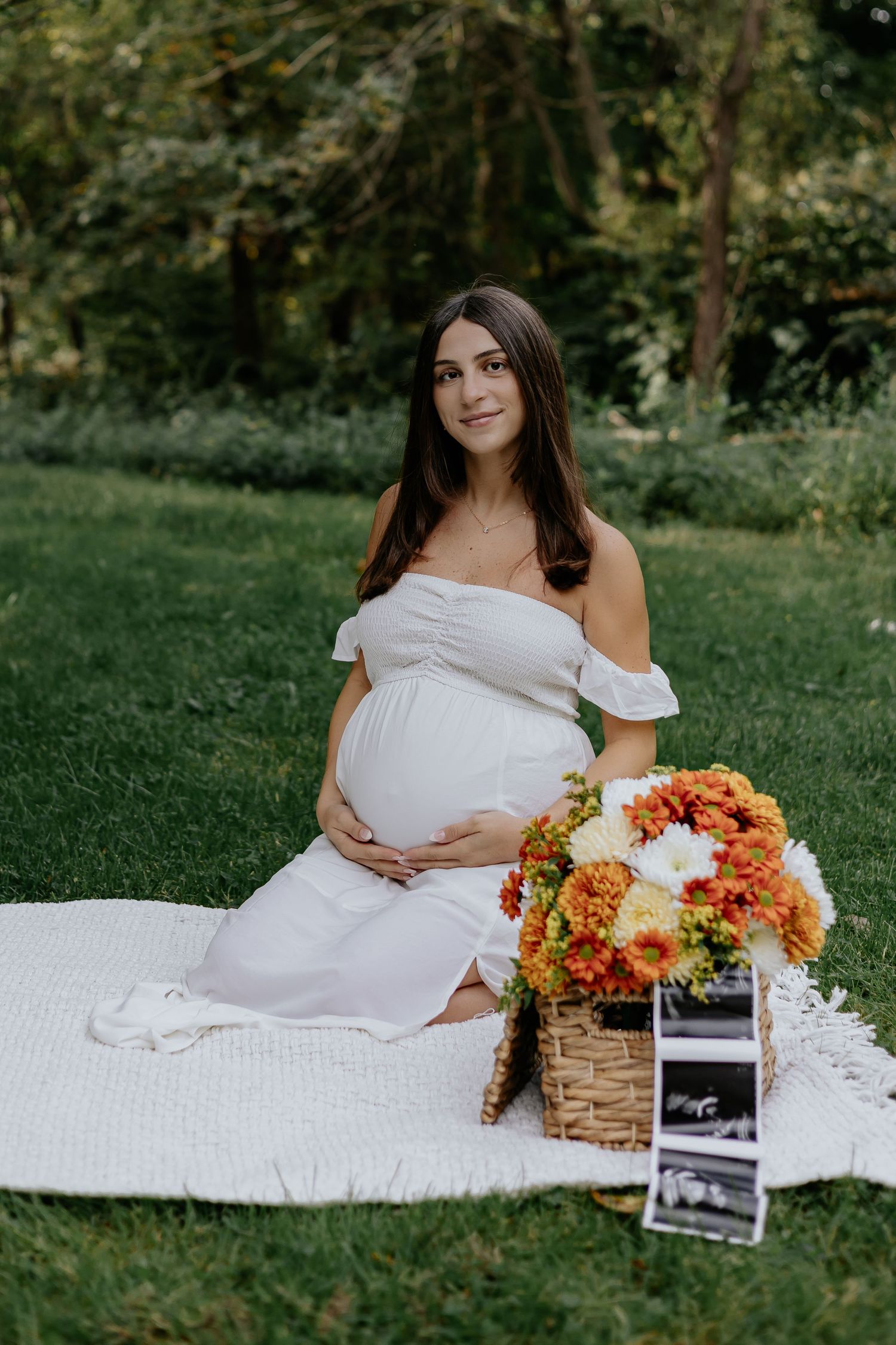 Pregnant woman in white off-shoulder dress sitting on blanket with fall flowers and ultrasound photos in park.