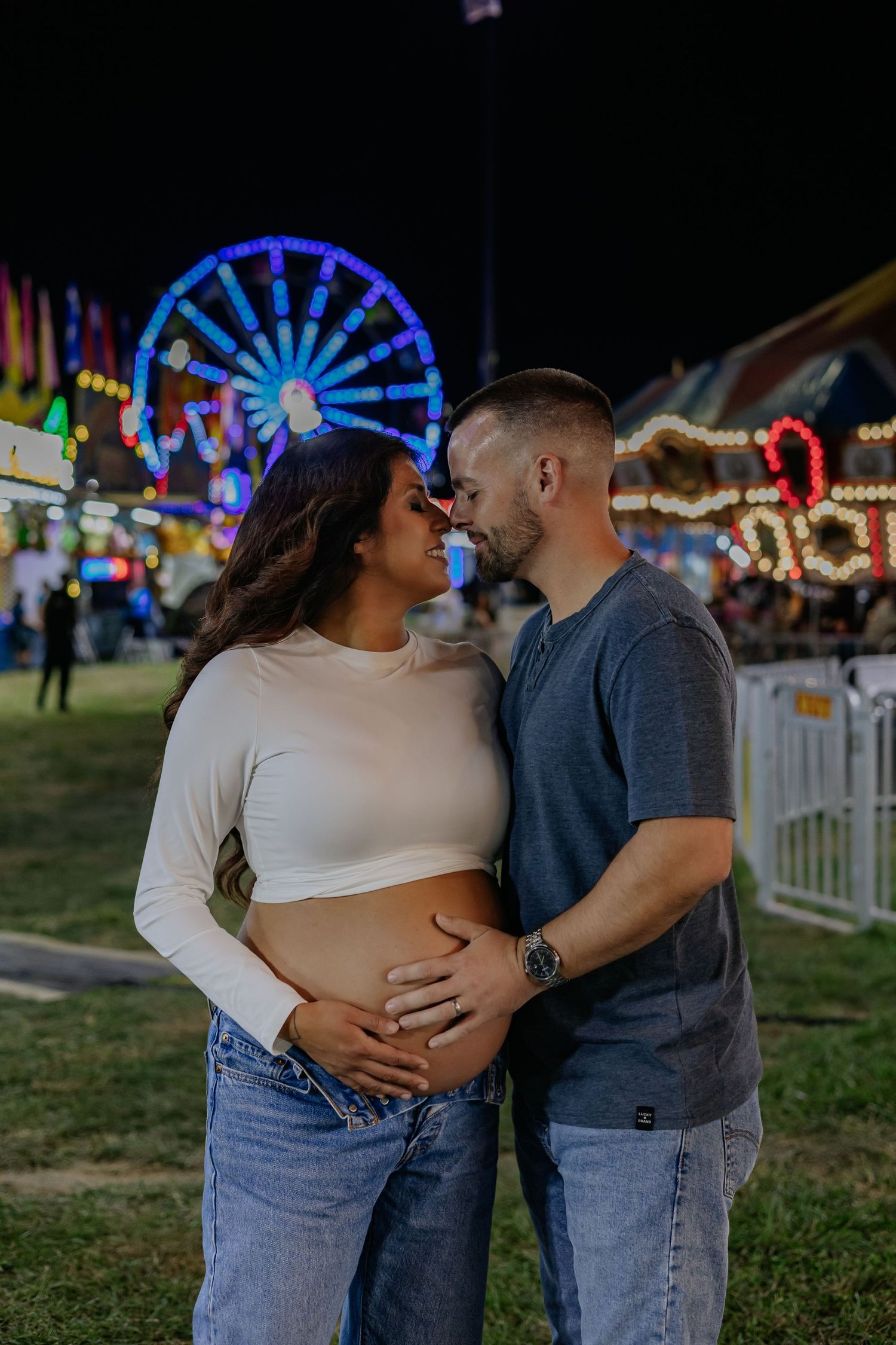Expecting couple tenderly holding baby bump with ferris wheel and carnival lights at night.