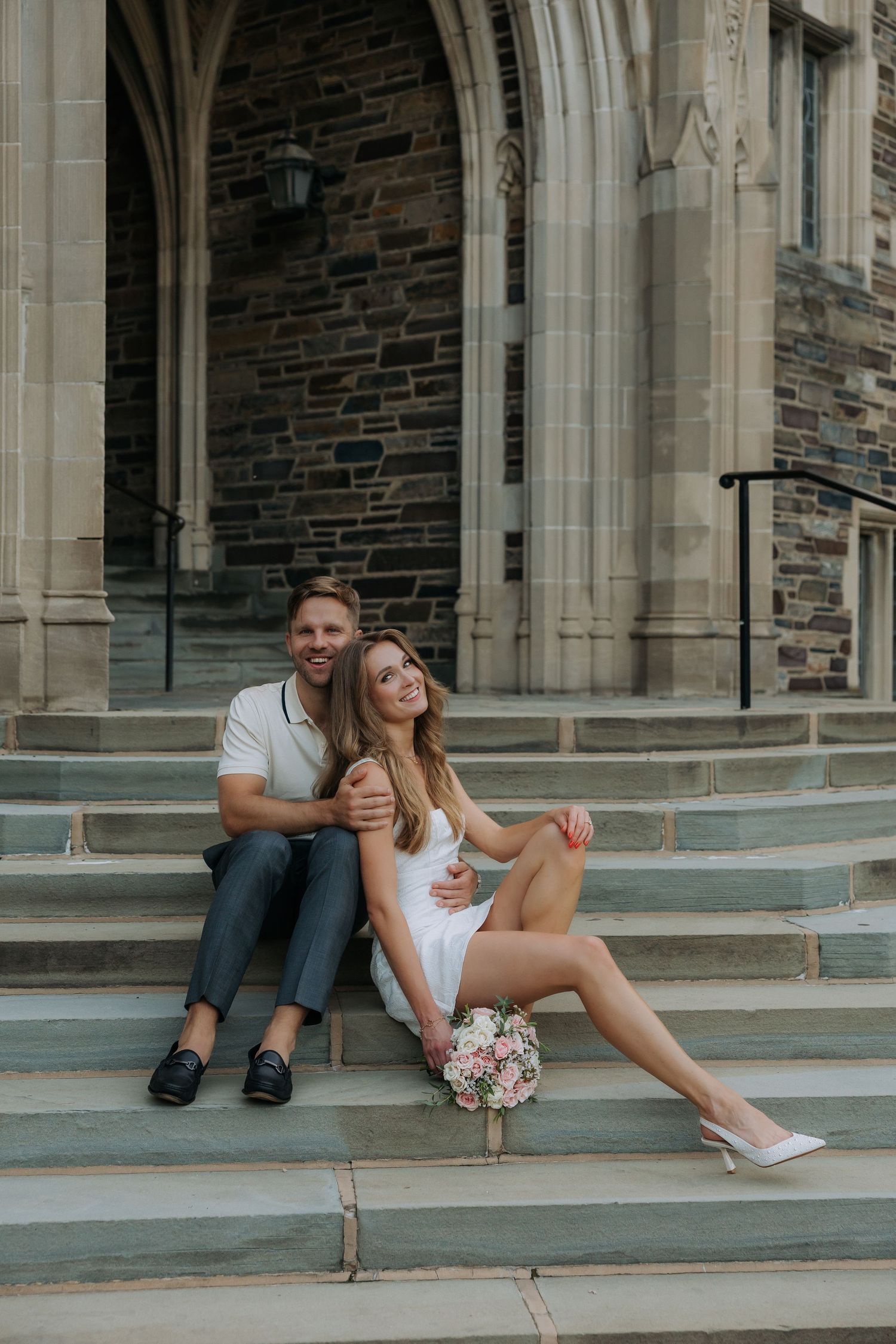 Couple sitting on stone church steps, woman holding a bouquet, smiling together.