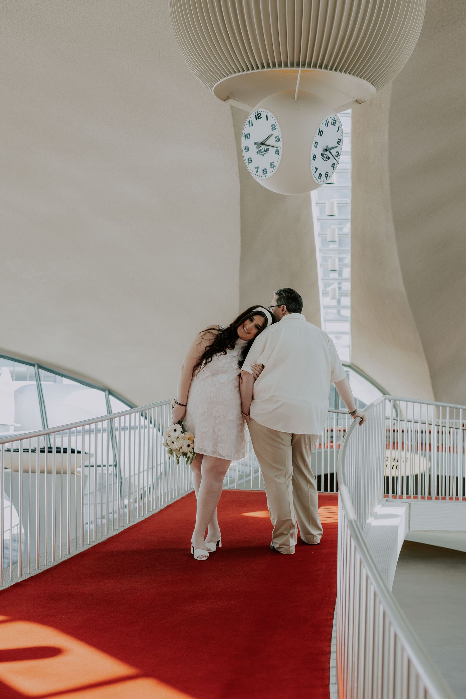 Pregnant bride in white dress holds bouquet on red carpet staircase with clock tower at TWA Hotel.