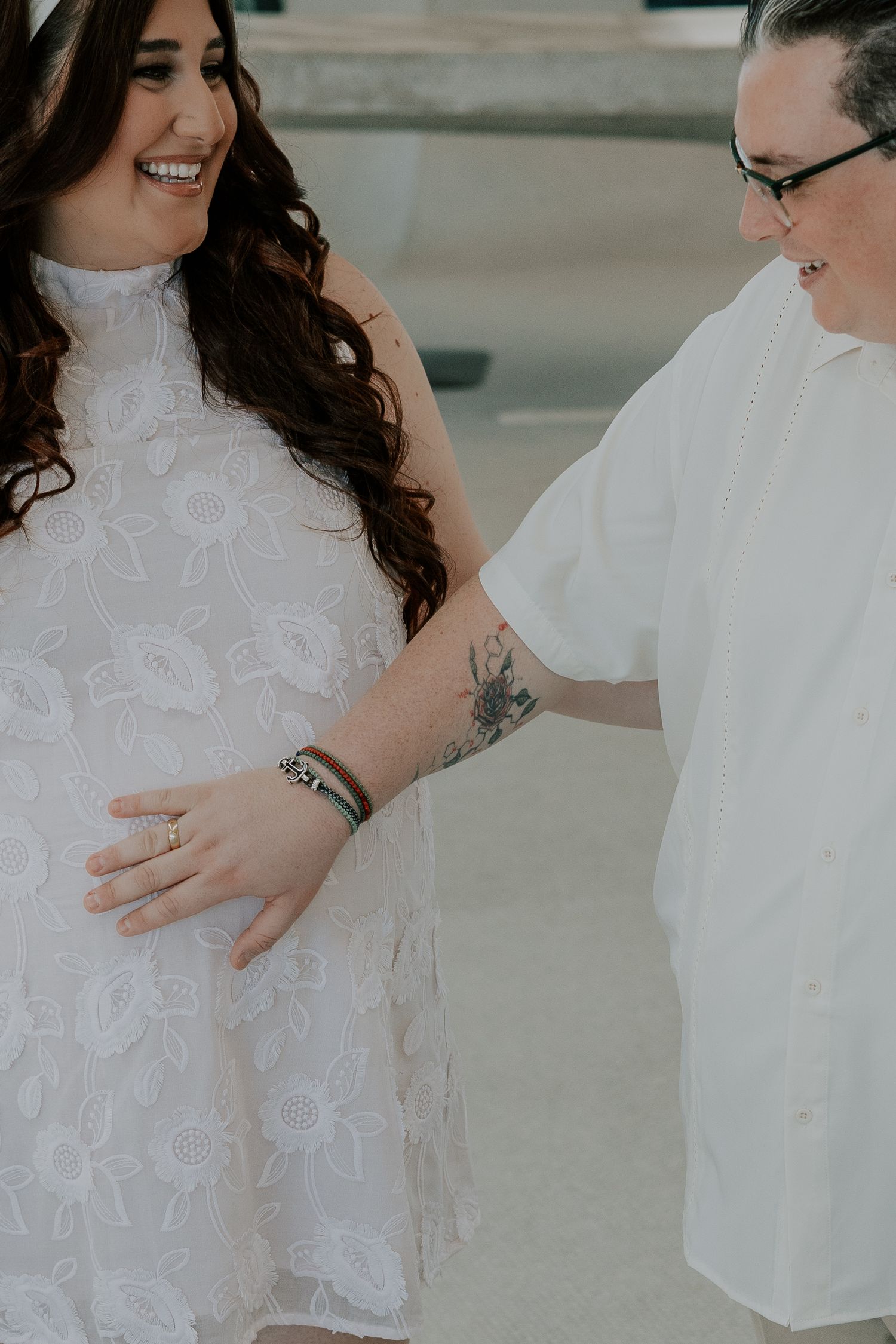 Smiling pregnant woman in white lace dress with partner gently touching her baby bump outdoors.