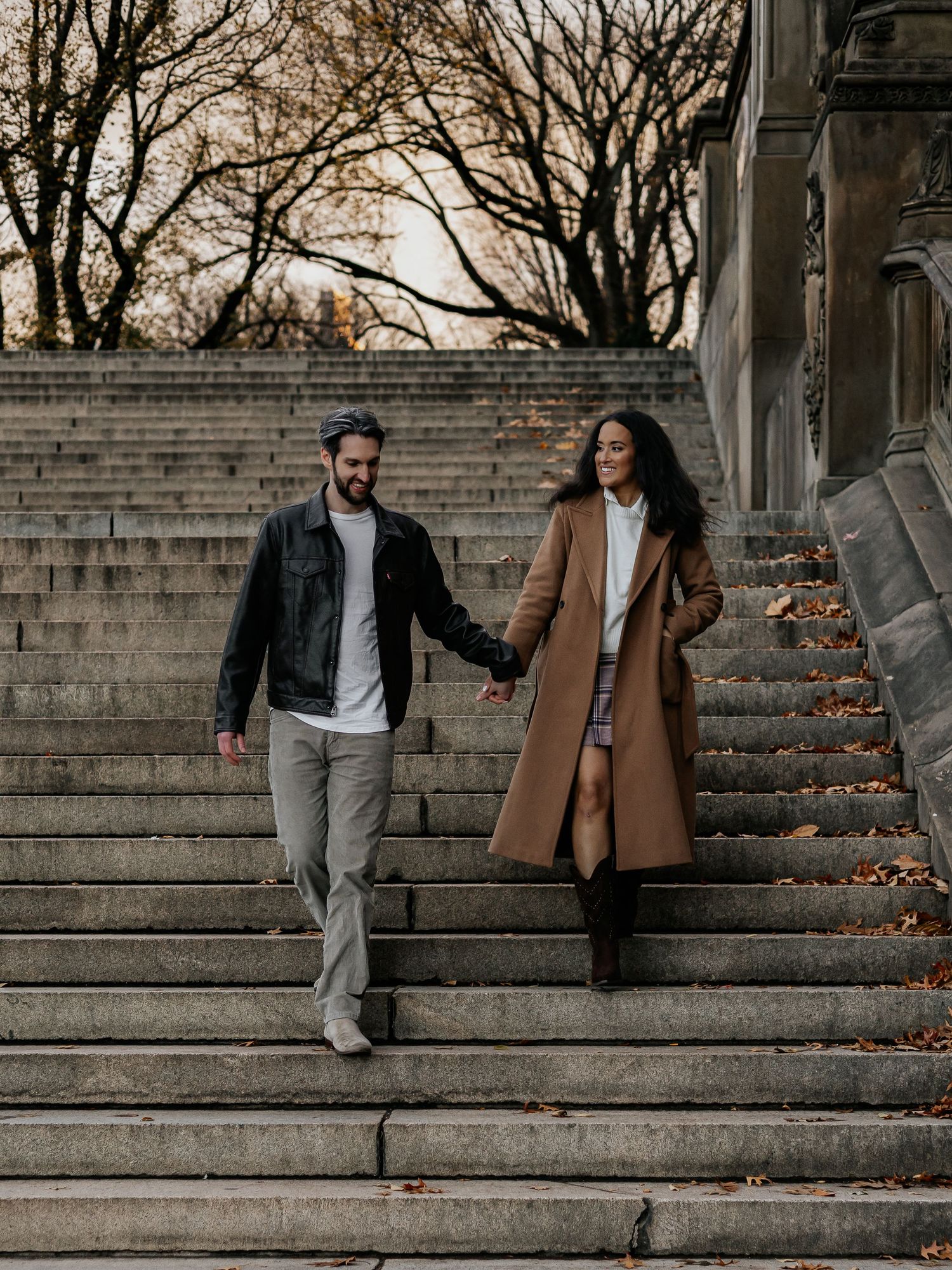 Happy couple holding hands descending Central Park steps surrounded by golden fall foliage.