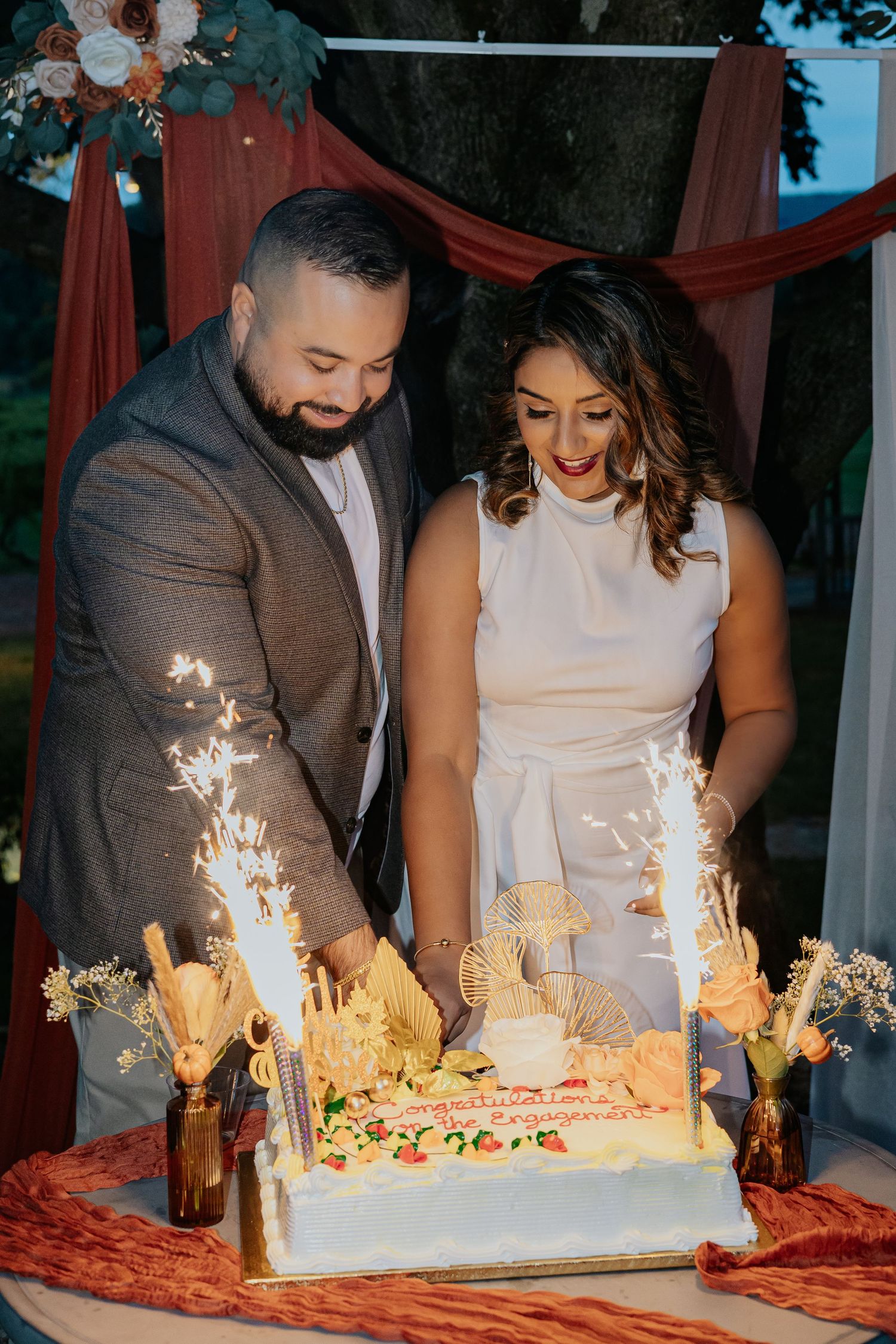 Couple cutting engagement cake with sparklers outdoors at dusk under floral arch.