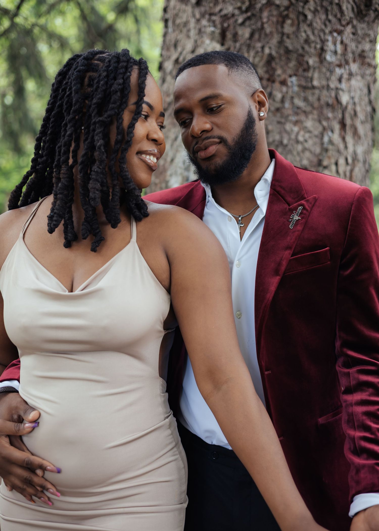 Romantic couple gazing at each other near tree, woman in champagne dress and man in burgundy velvet blazer.