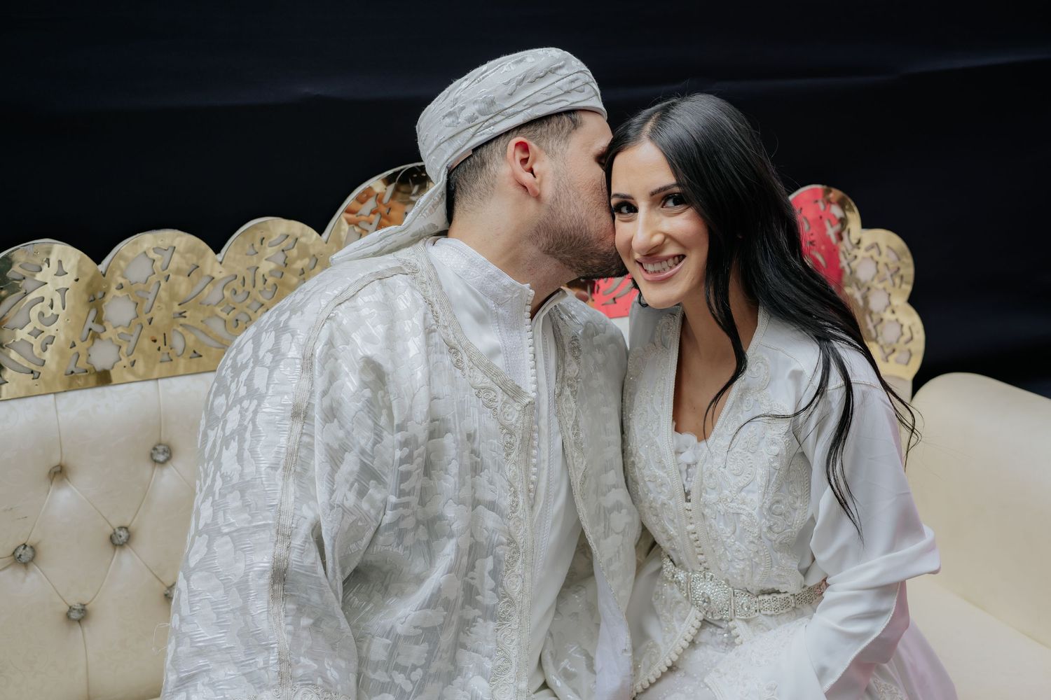 Couple in white traditional Moroccan attire share an intimate moment seated on ornate gold wedding sofa.