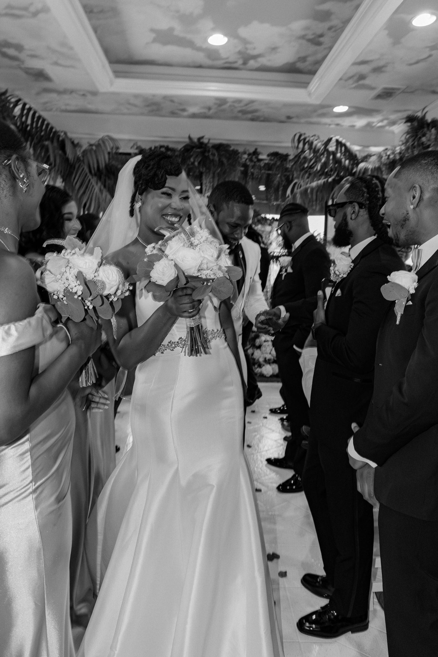 Black and white photo of a radiant bride holding a bouquet while walking the aisle flanked by bridesmaids and groomsmen.
