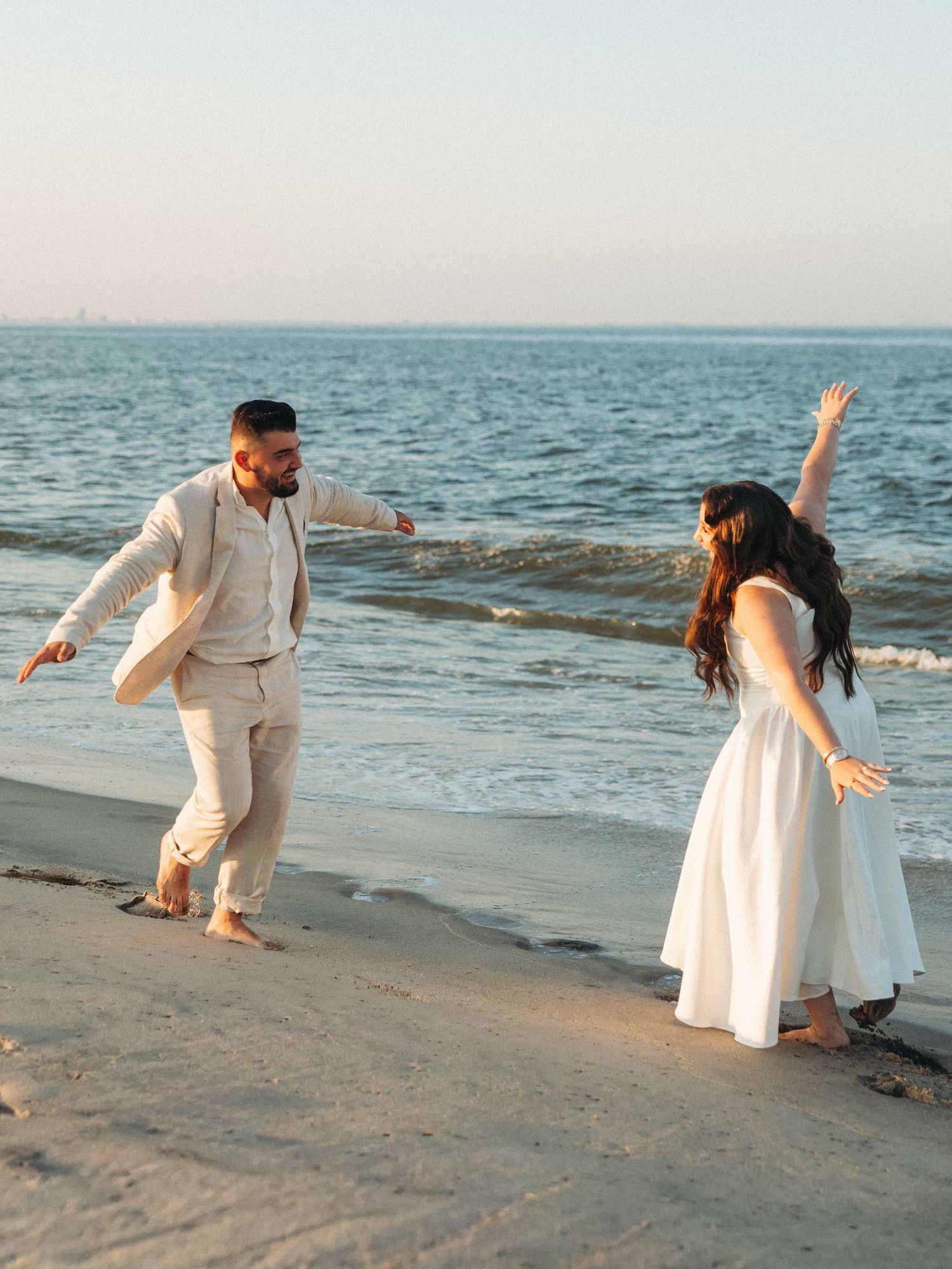A couple dances barefoot on a sandy beach at sunset, wearing white and beige outfits with ocean waves behind them.