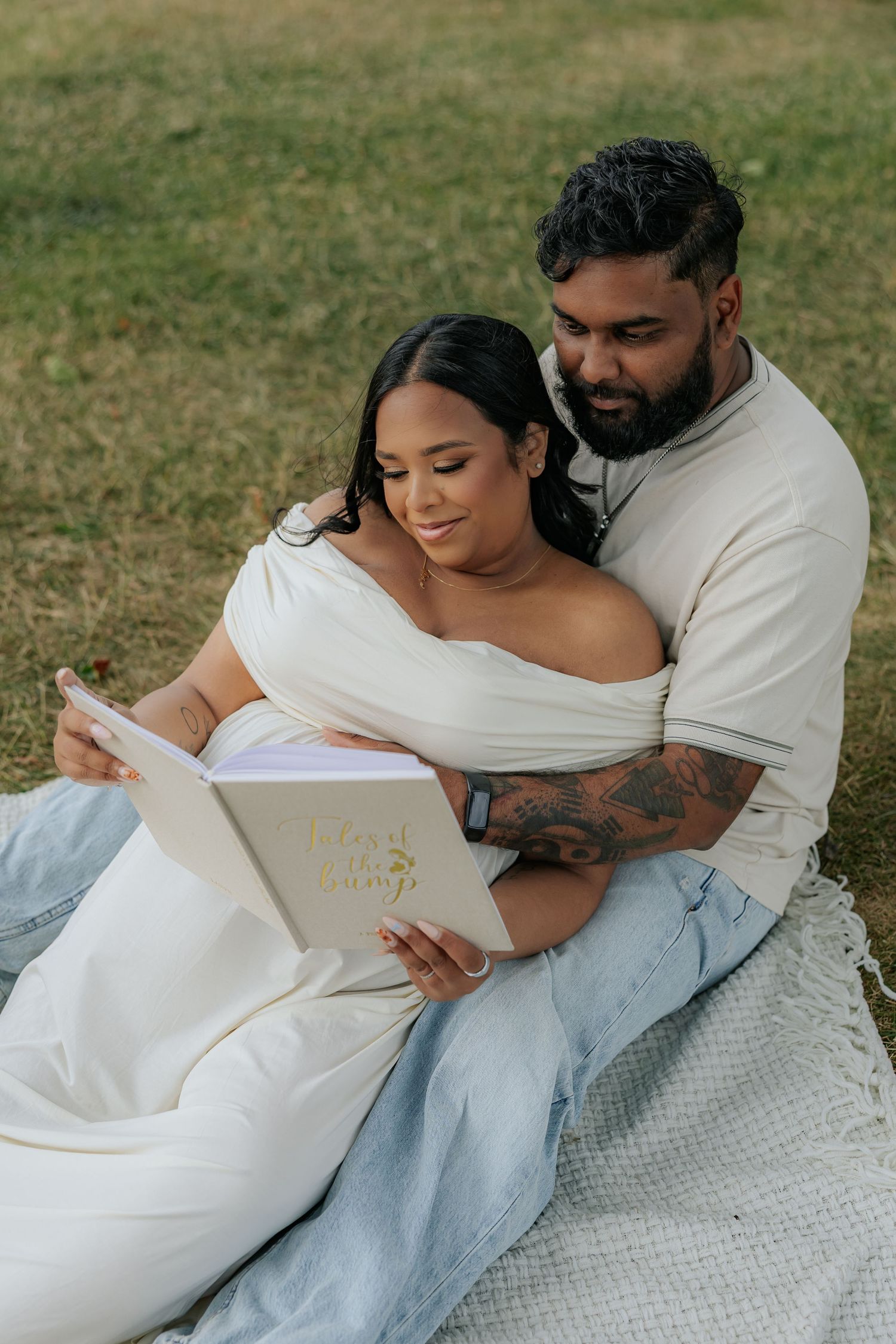 Couple sitting on blanket in grass, woman in white off-shoulder dress reading a baby book together during maternity photoshoot.