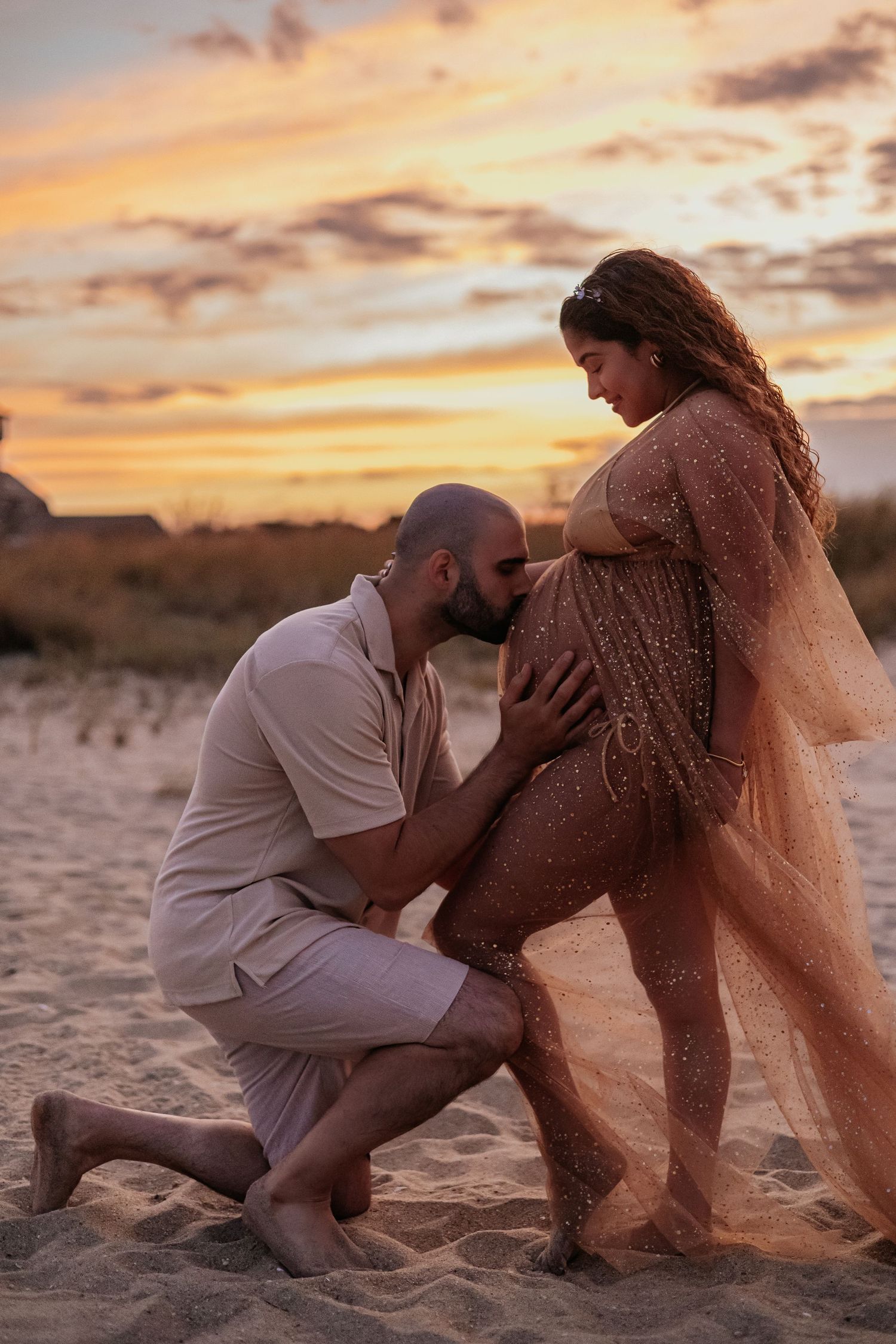 Man kneeling to kiss pregnant belly on sandy beach during vibrant orange sunset maternity session.