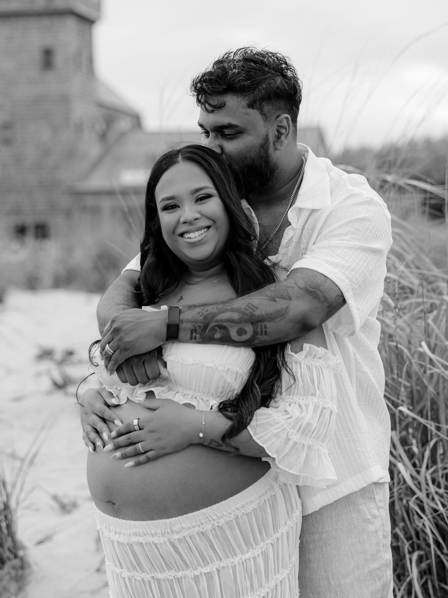Couple embracing on beach during maternity photo shoot, man kissing woman's head while cradling her baby bump.