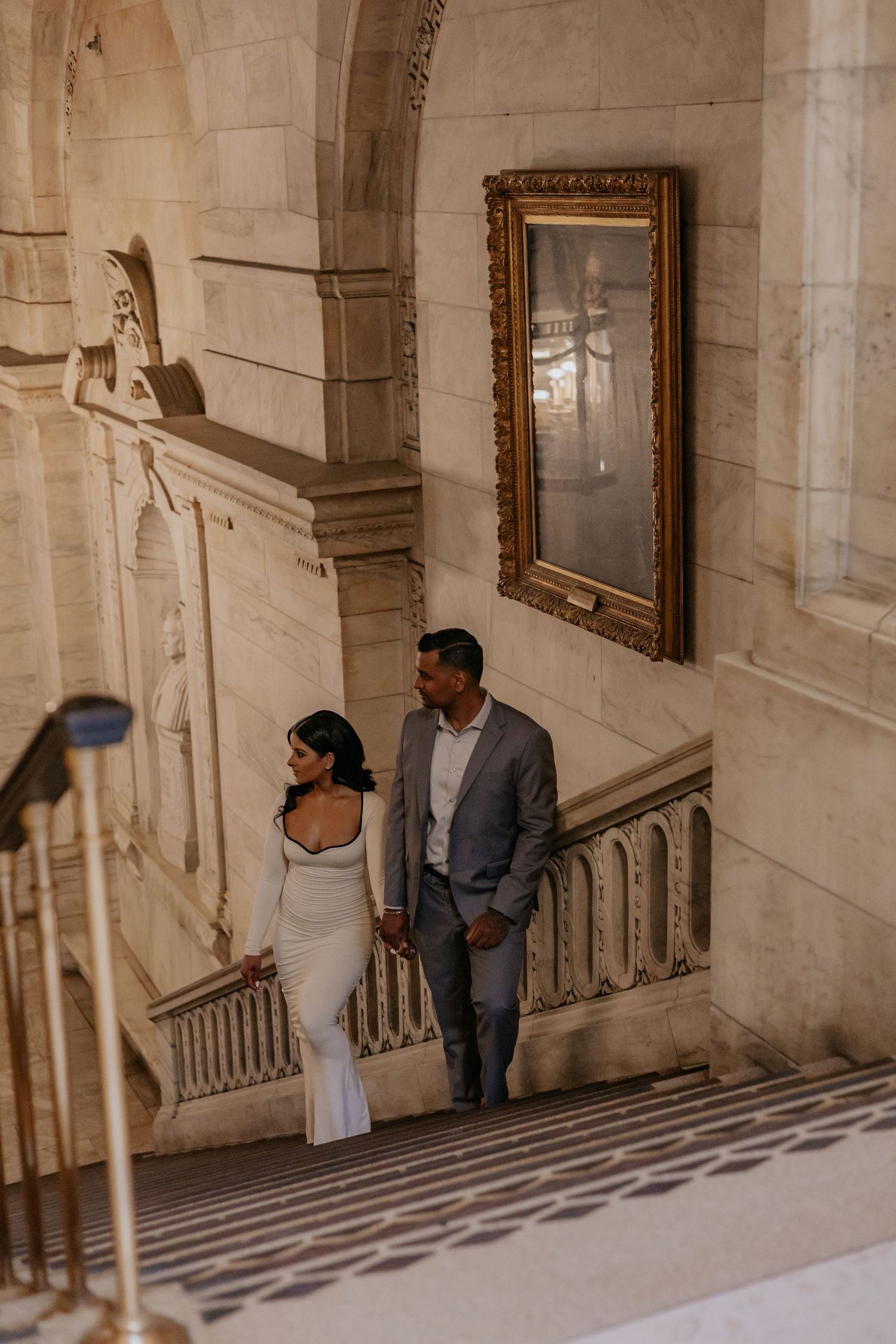 Couple holds hands walking past gold-framed portrait along marble archway wall.
