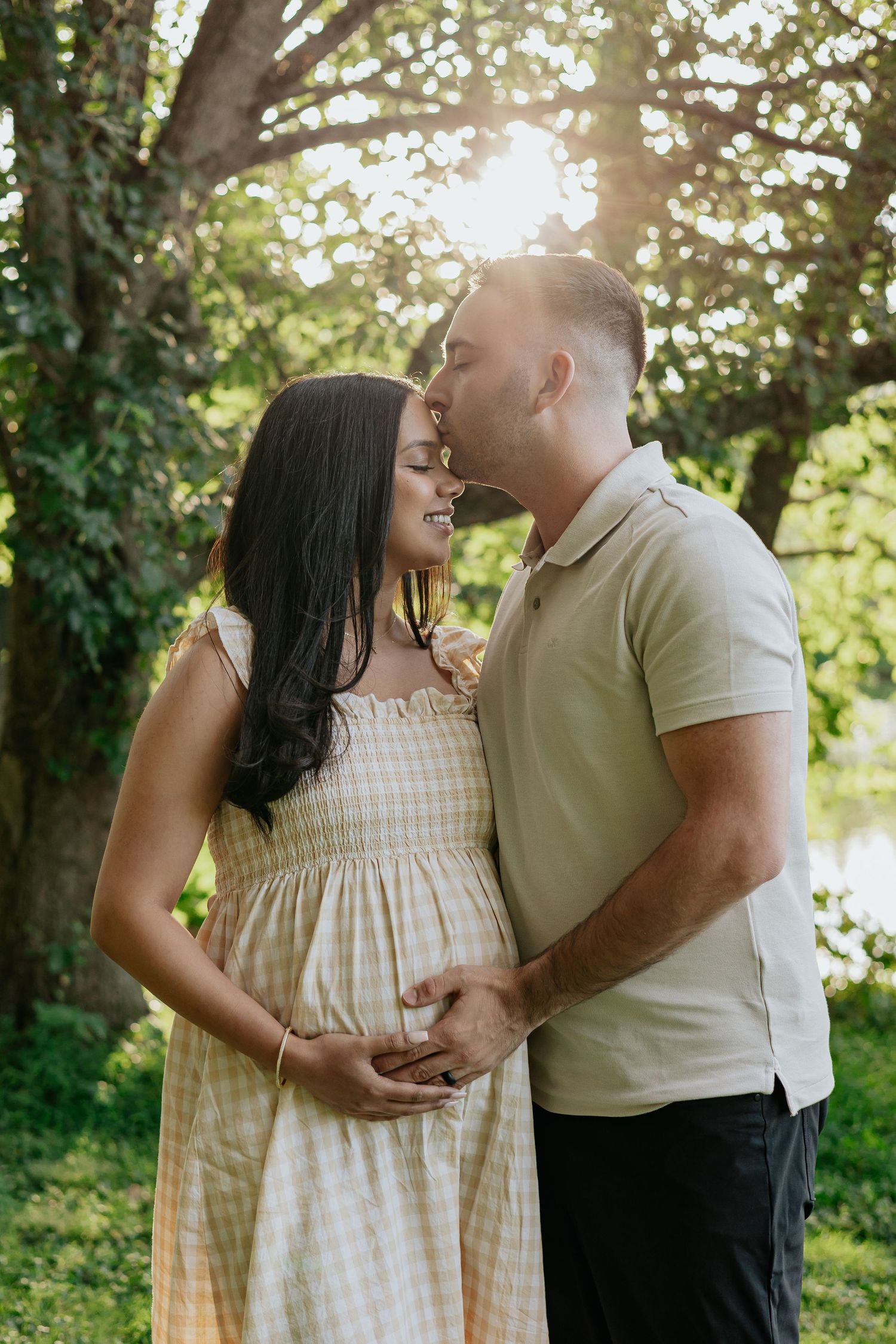 Couple sharing tender moment outdoors, man kissing pregnant woman's forehead while holding her baby bump in golden sunlight.