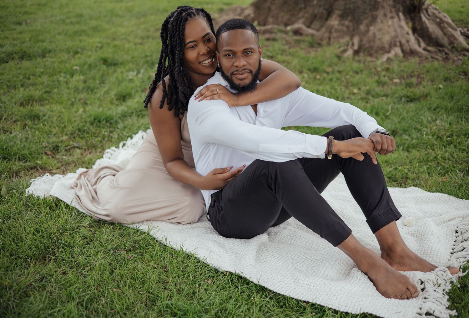 Smiling Black couple sitting together on white blanket in green park, posing for engagement photos.