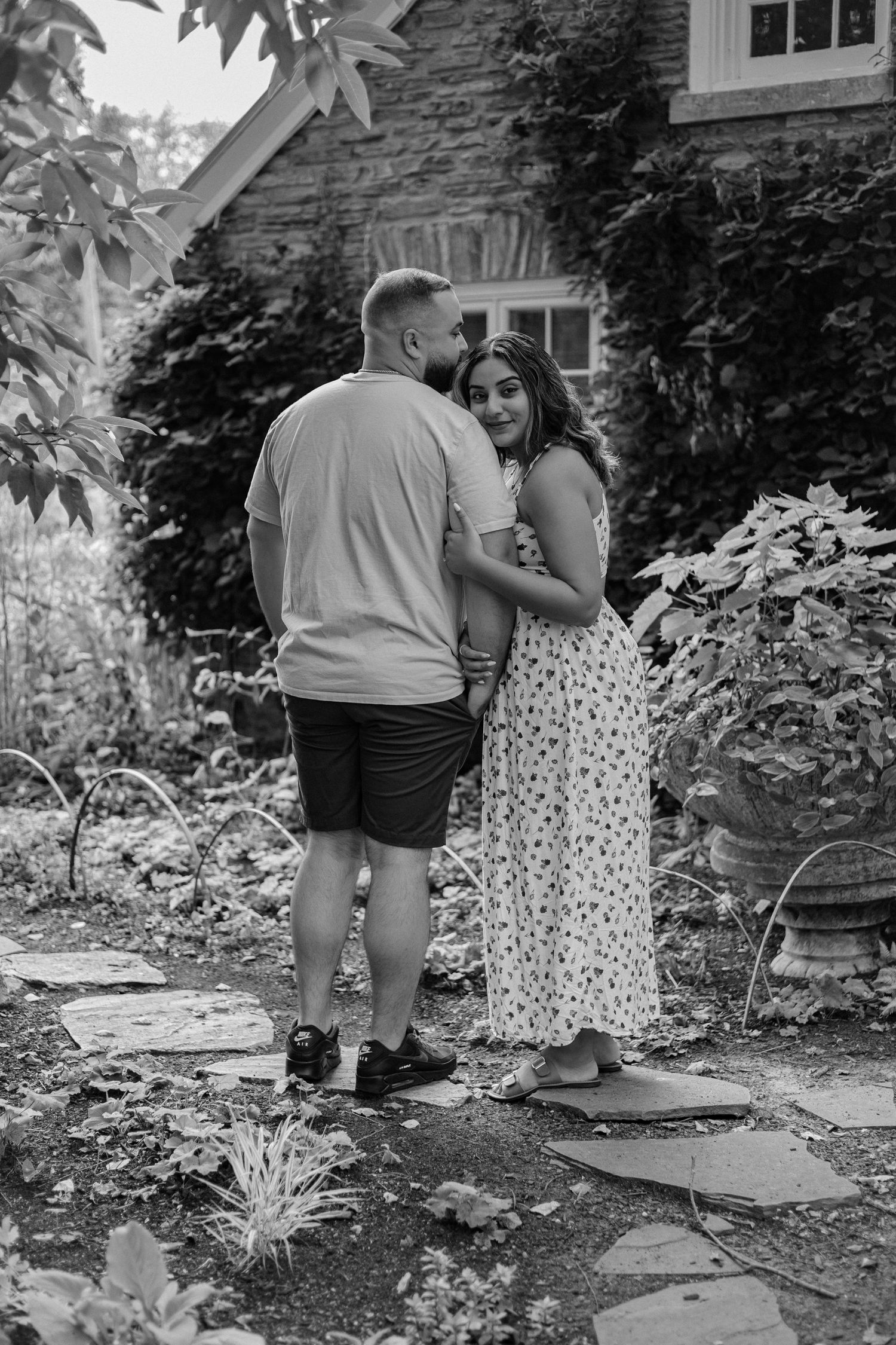 Black and white photo of a couple embracing in a lush garden beside a stone cottage.