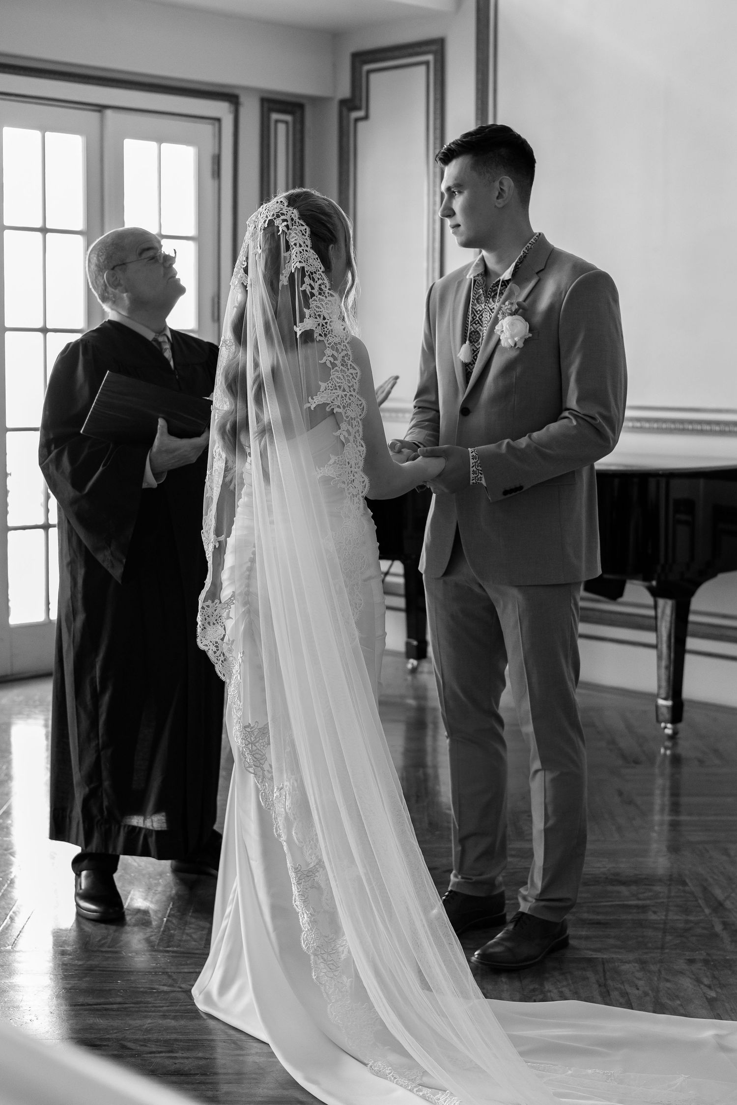 Black and white photo of bride and groom exchanging vows with officiant near grand piano indoors.