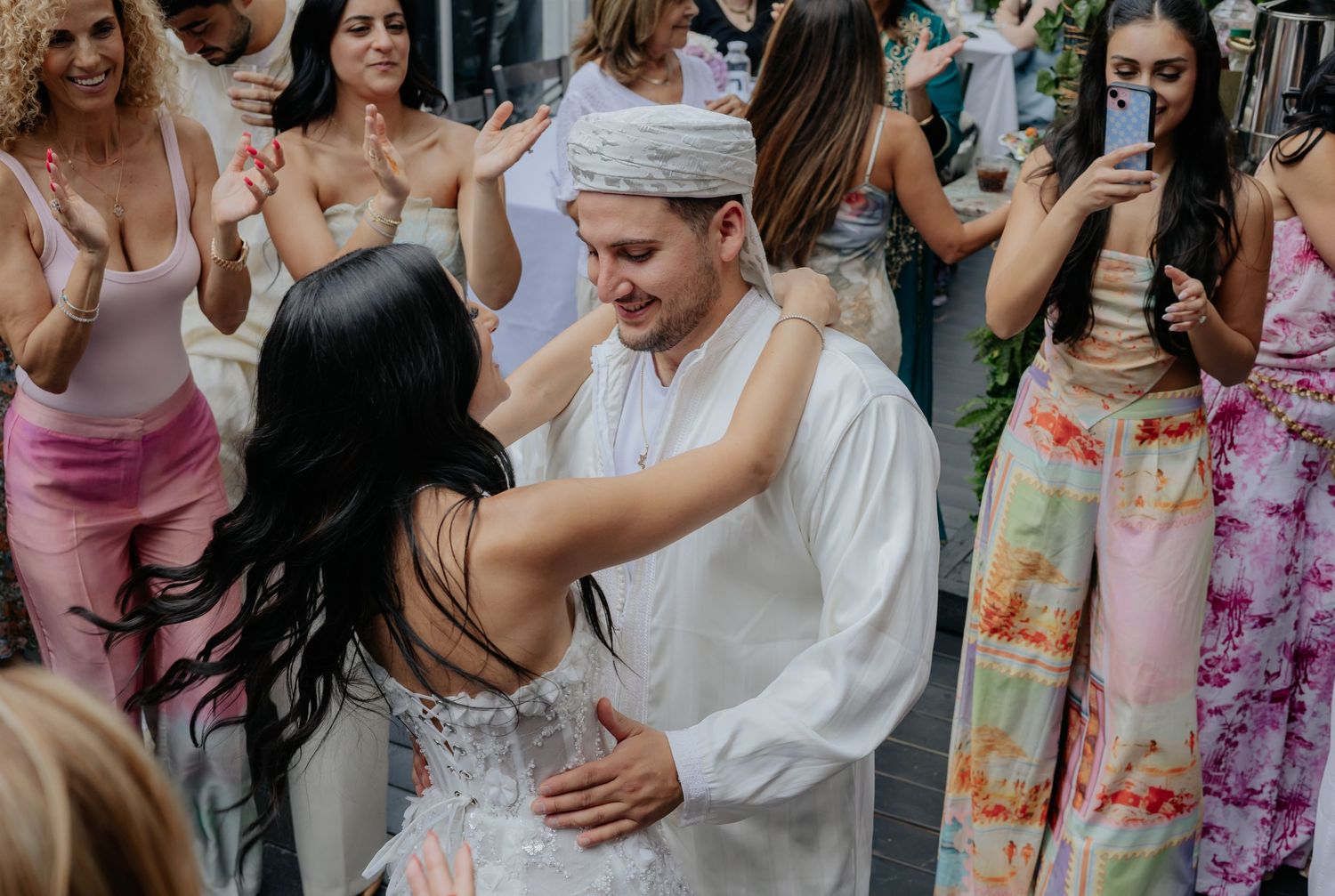 Bride and groom in white traditional outfits share a first dance surrounded by celebrating wedding guests.