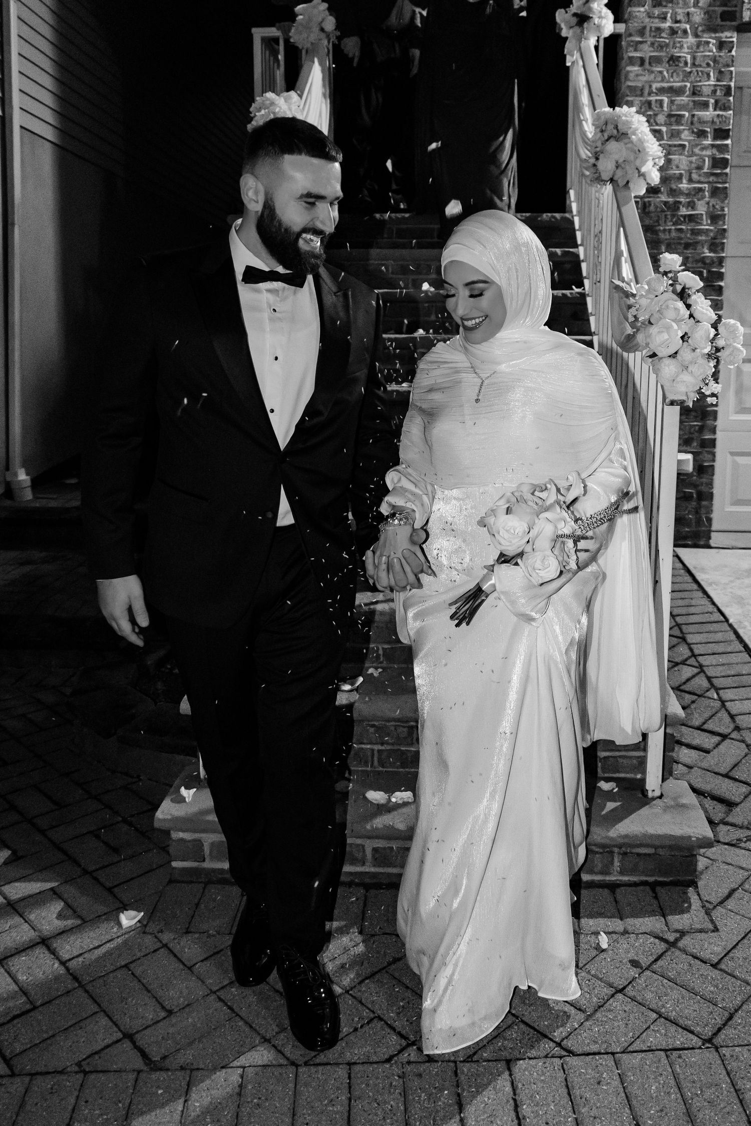 Black and white photo of Muslim bride in hijab and groom in tuxedo walking together at wedding.