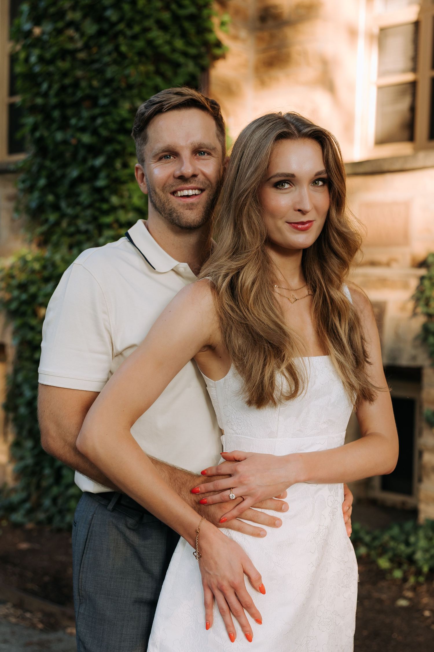 Smiling couple embracing outdoors near ivy-covered wall, woman in white dress with long wavy hair, man in polo shirt.