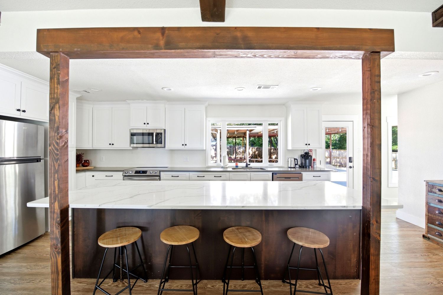 Modern kitchen with marble island, wood bar stools, white cabinets, and stainless steel appliances framed by rustic wood beams.
